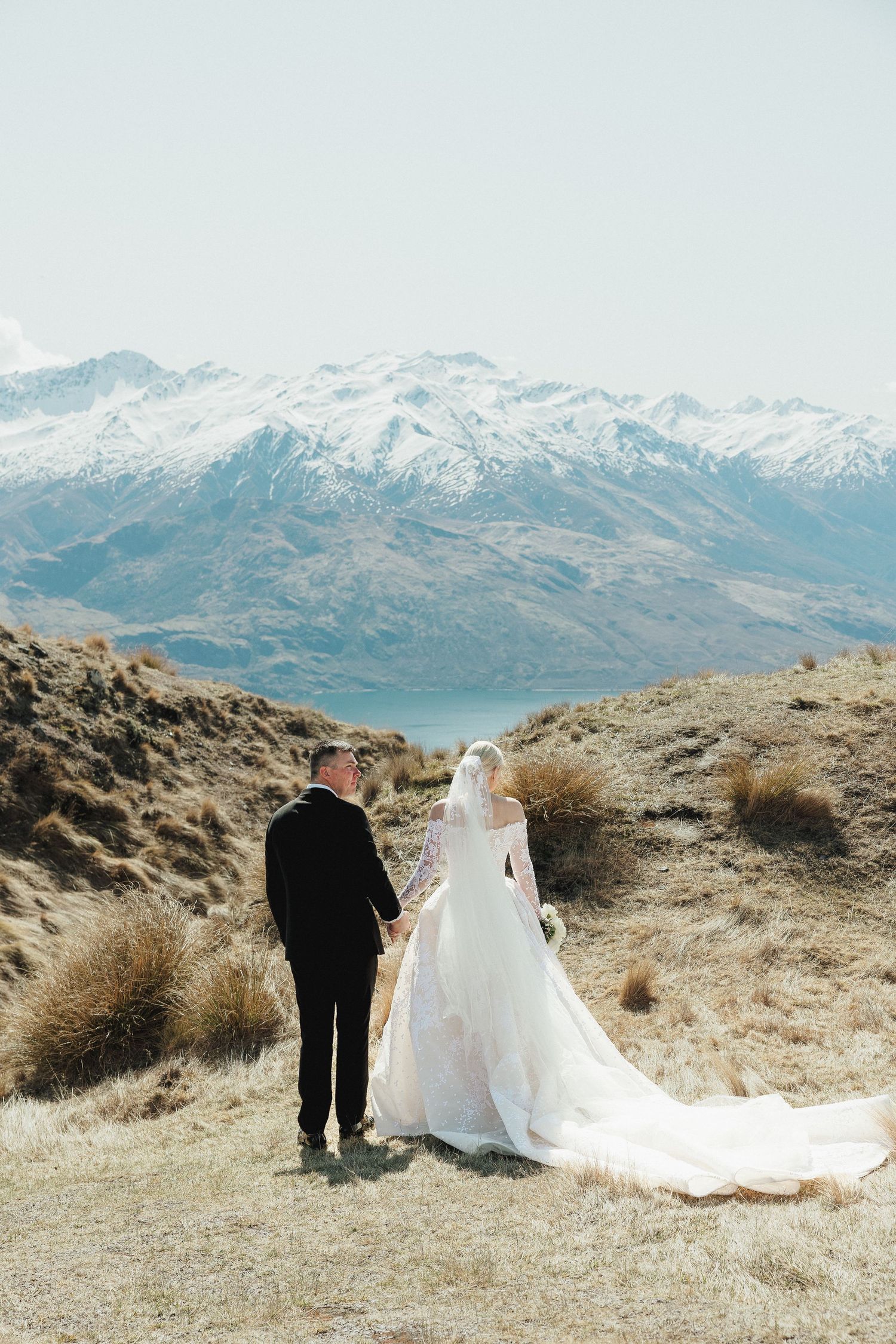 Bride and groom walk hand in hand through alpine meadow with snow-capped mountains in Queenstown, New Zealand.