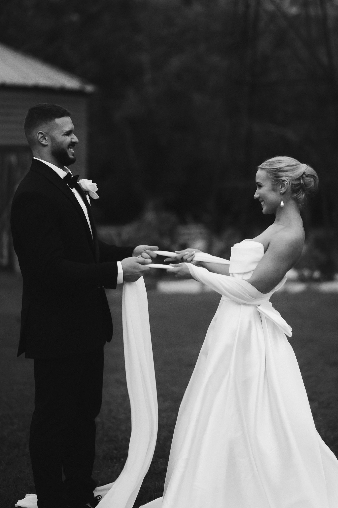Black and white photo of smiling bride and groom exchanging vows outdoors, bride in strapless gown with bow, groom in tuxedo.
