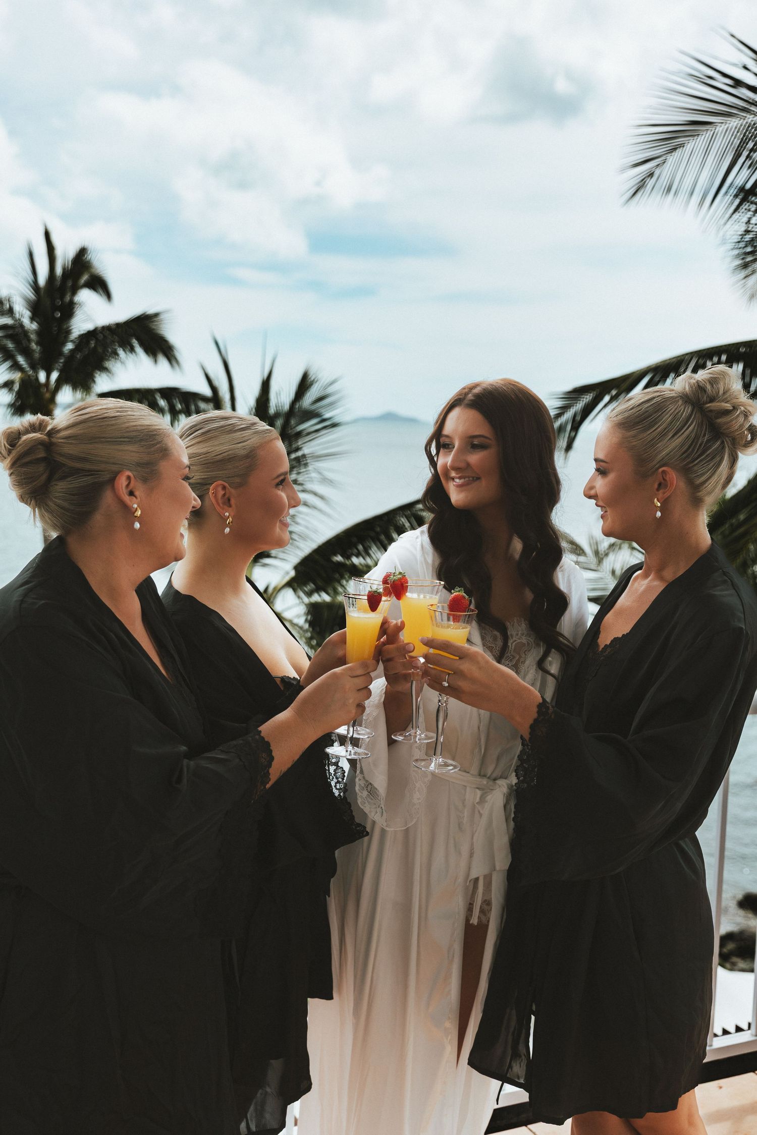 Bridesmaids in black robes toasting mimosas with bride in white robe at tropical beach resort in Whitsundays, Queensland.