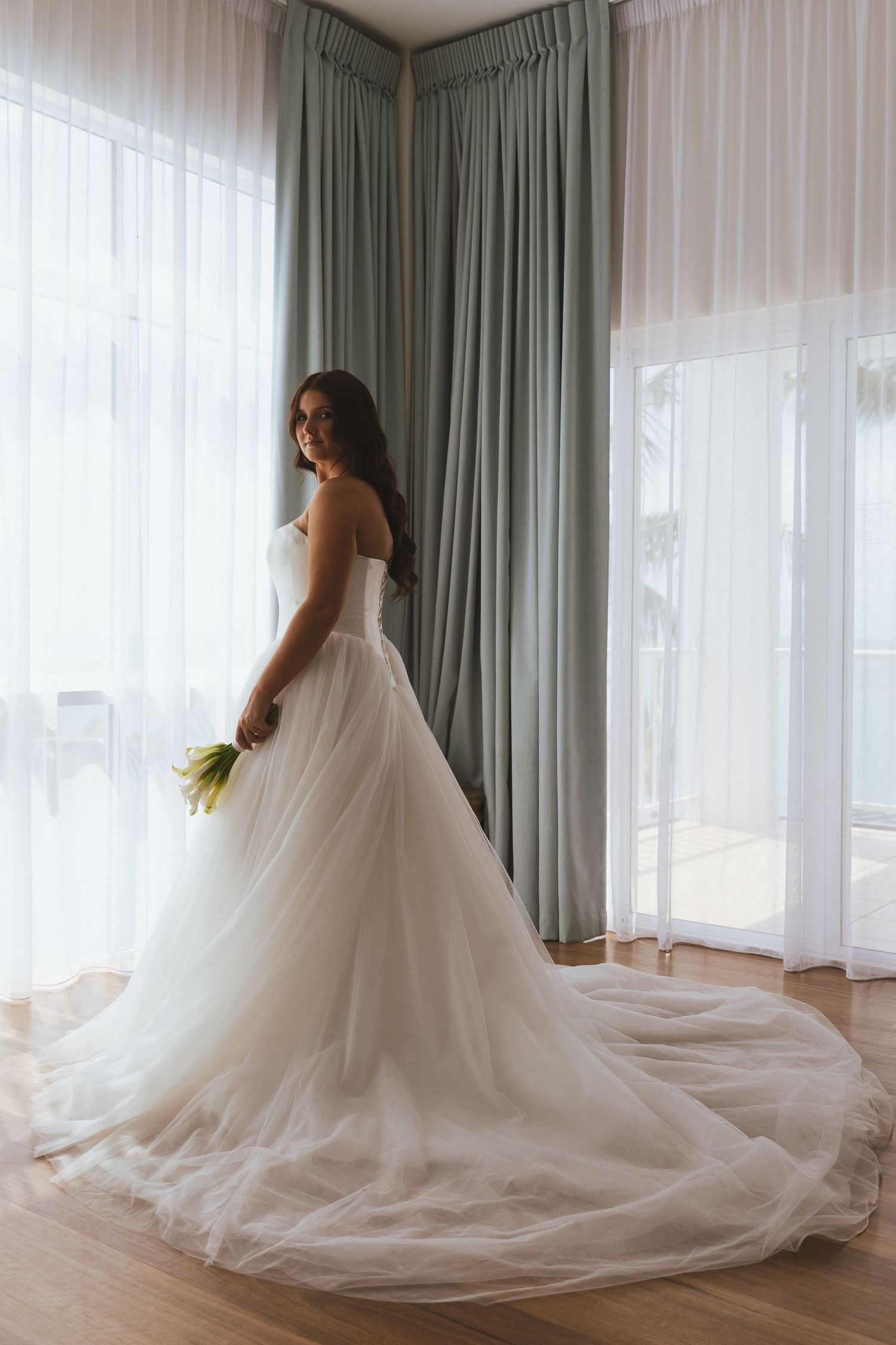 Elegant bride in strapless ball gown with long tulle train standing by floor-length curtained windows.