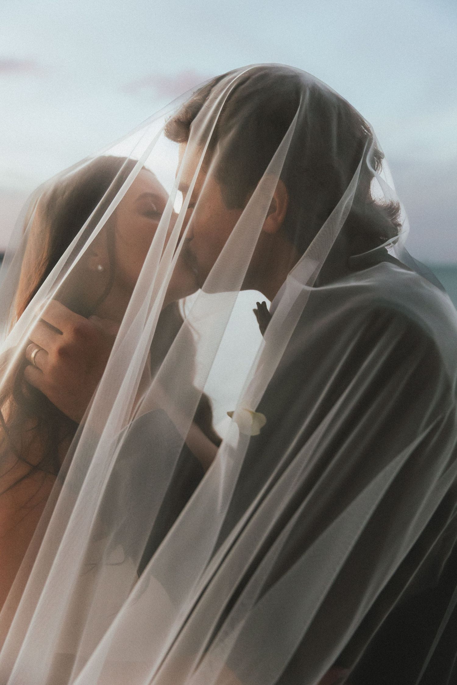 Bride and groom sharing an intimate kiss beneath a flowing sheer wedding veil with soft sky backdrop.
