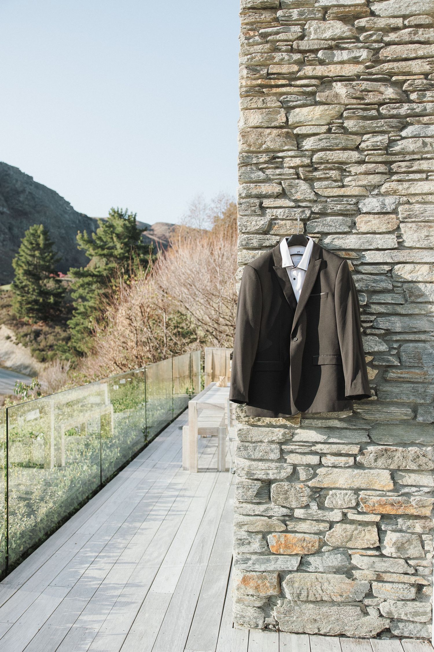 Grooms dark suit jacket hanging on stone wall of mountain venue balcony with scenic valley view.