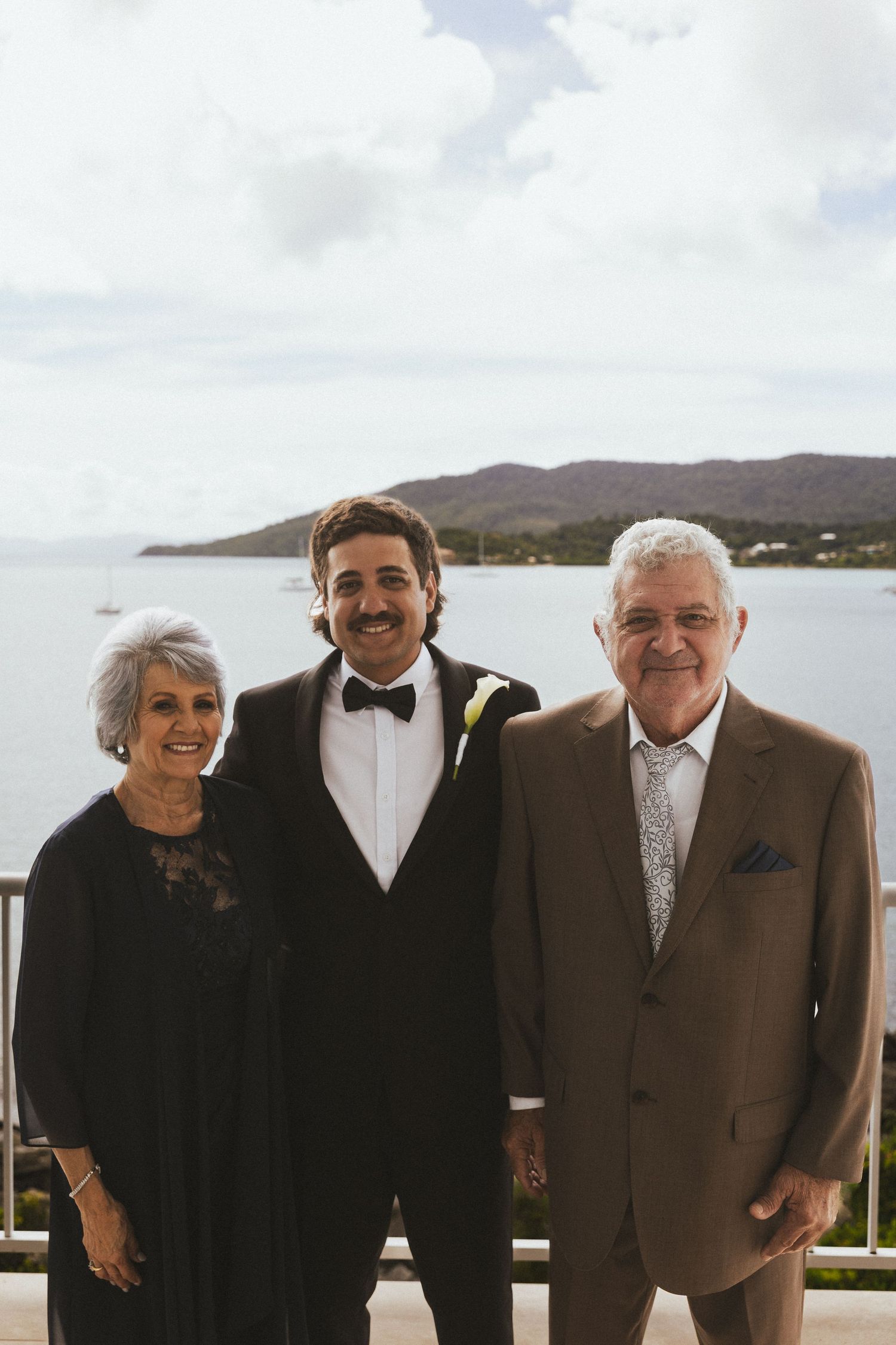 Groom in black tuxedo posing with proud parents against scenic waterfront and mountain backdrop in Whitsundays, Queensland.