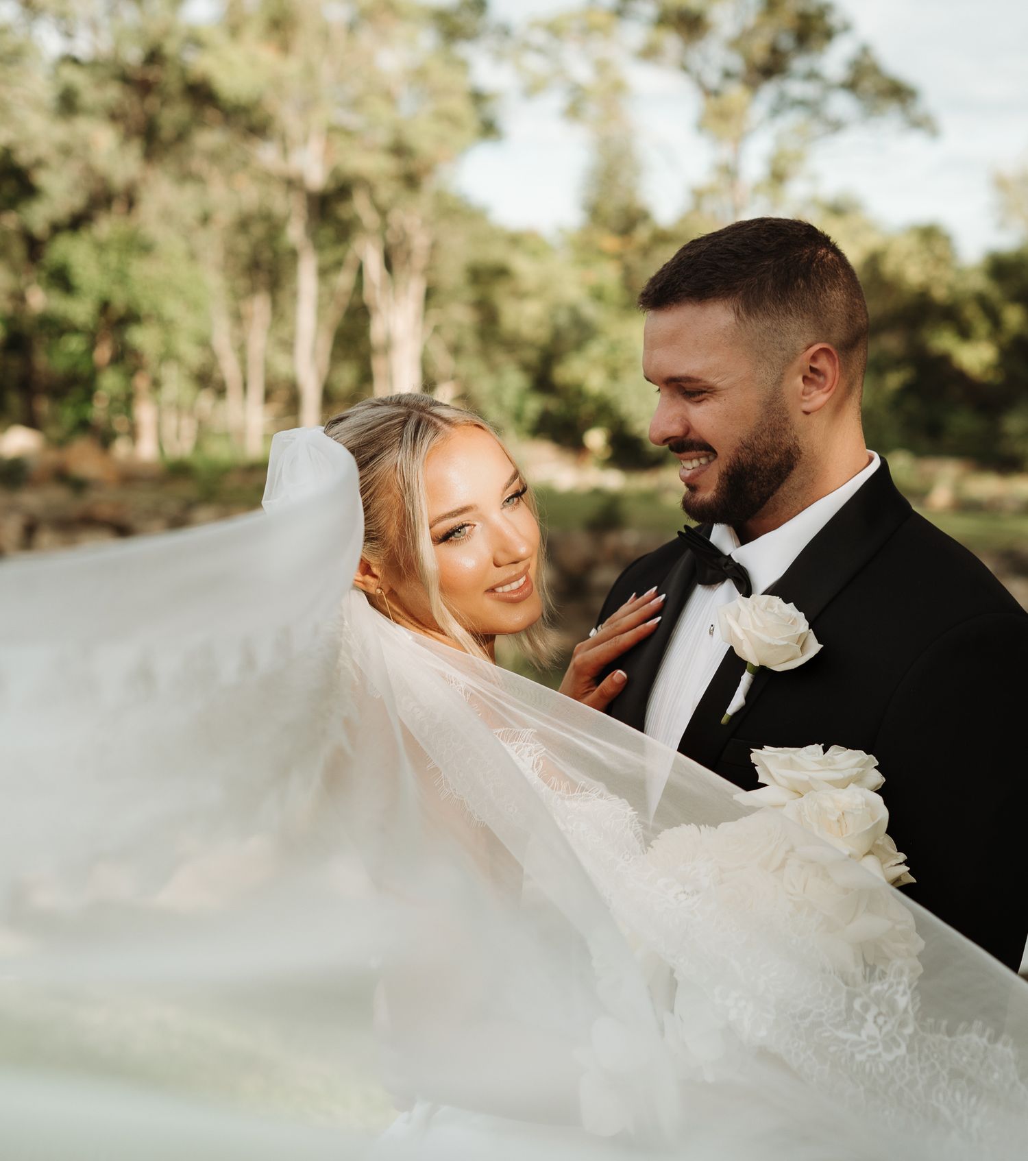 Bride and groom sharing a romantic moment outdoors, her white veil flowing in the breeze as they smile at each other.