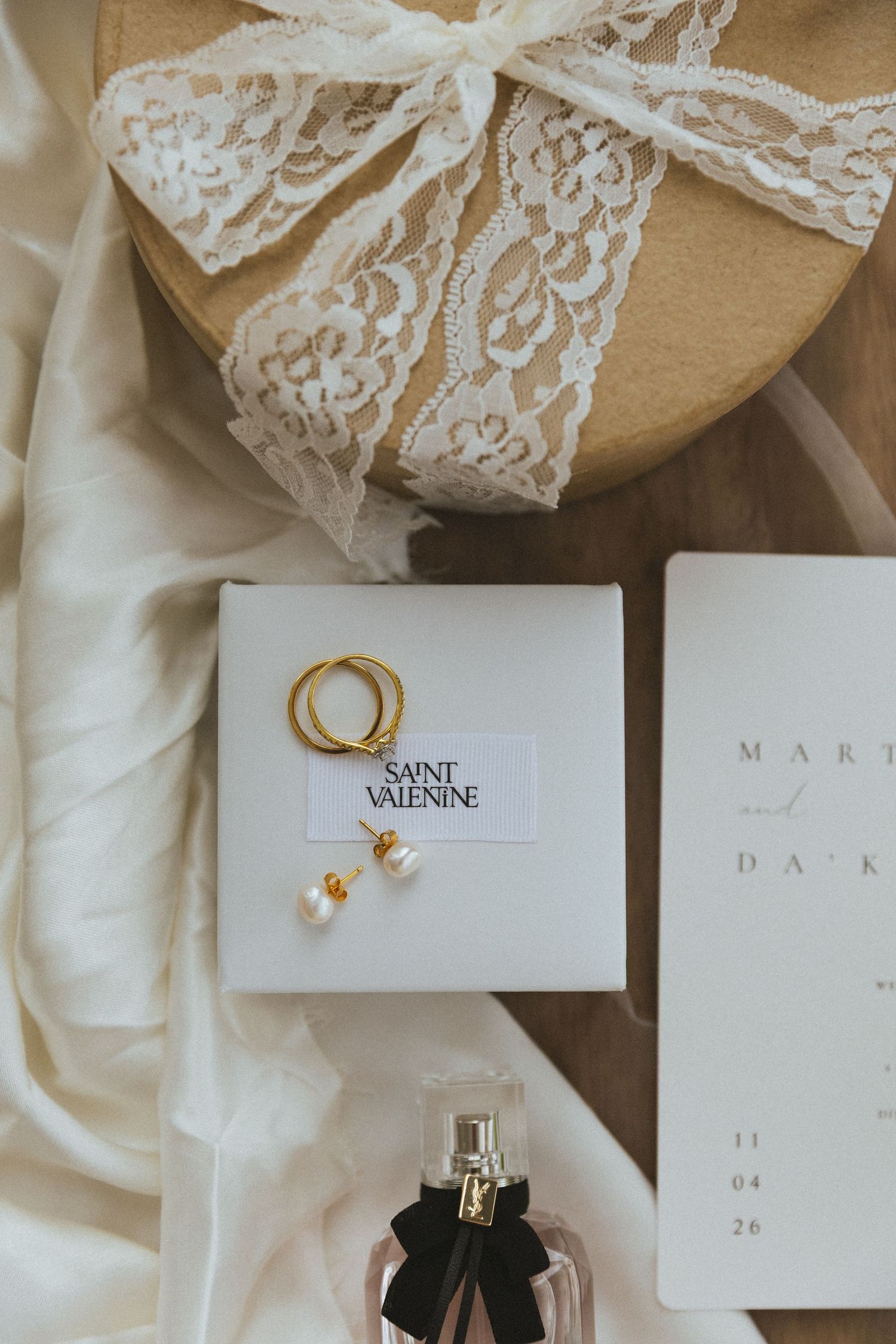 Elegant wedding flat lay featuring Saint Valentine gold rings, pearl earrings, perfume, and lace gift box.