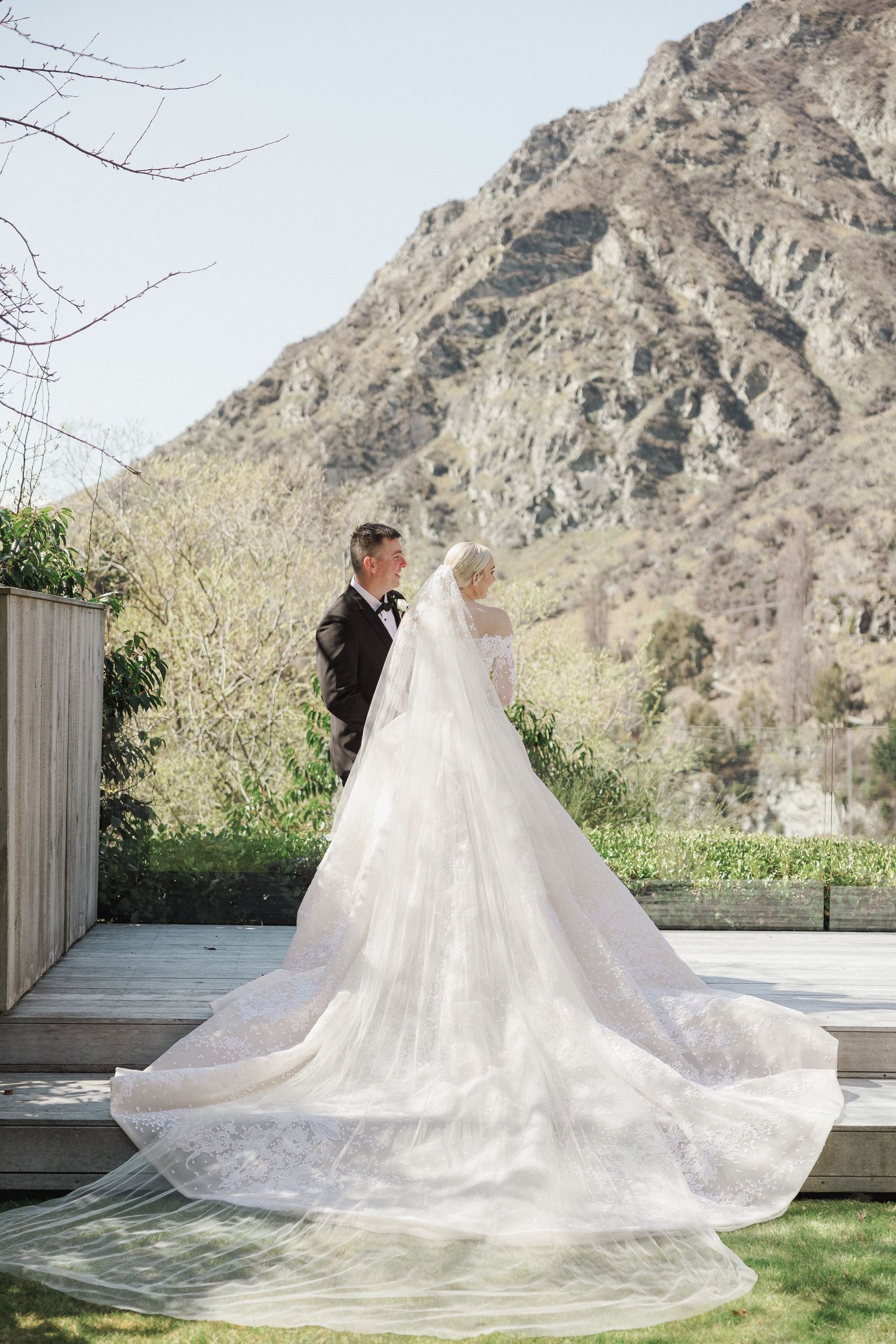 Bride and groom stand together showcasing stunning cathedral lace veil train with dramatic mountain backdrop in Queenstown, New Zealand.