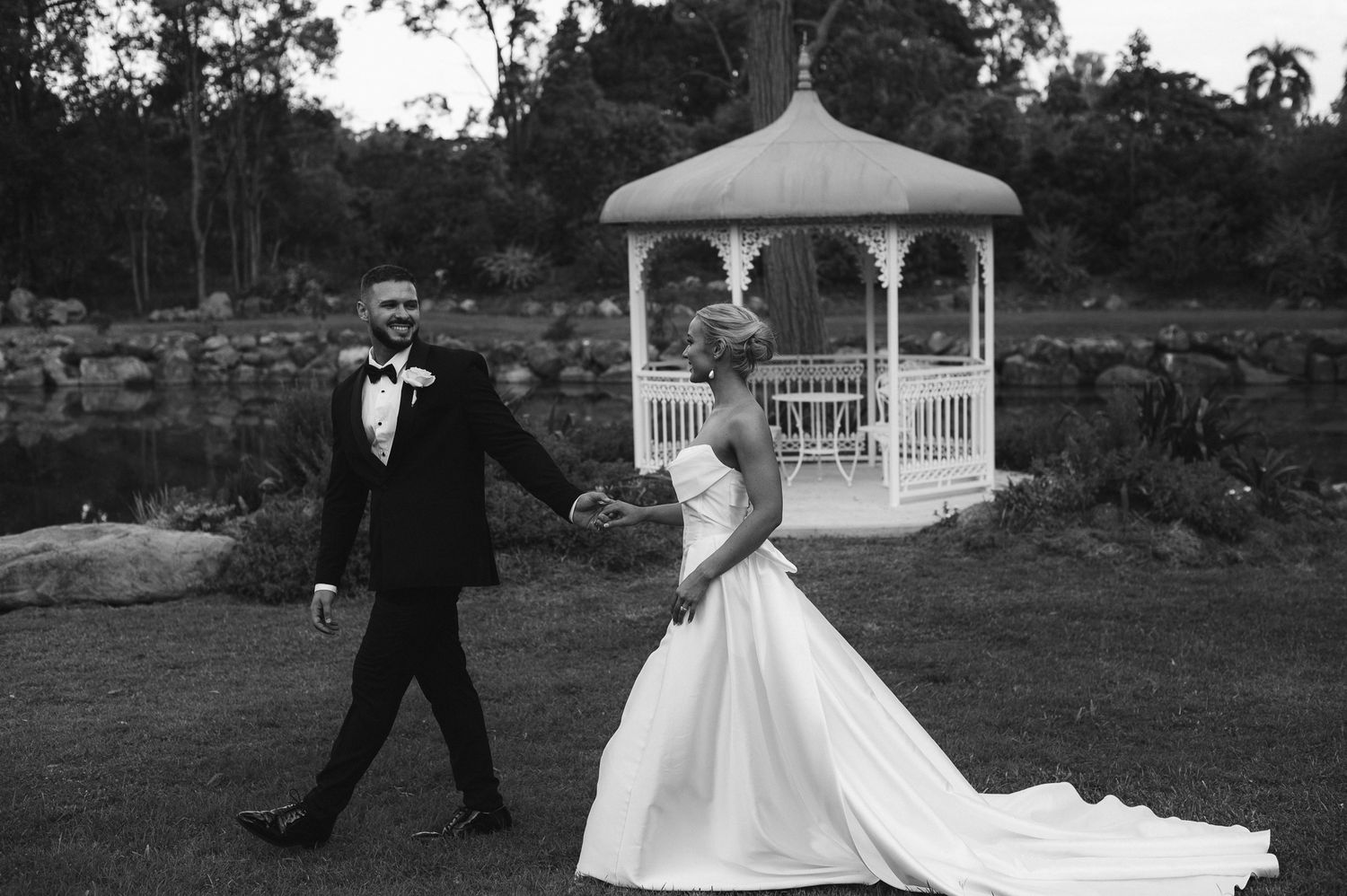 Bride and groom walk hand in hand near a white gazebo by a pond in a black and white wedding photo.