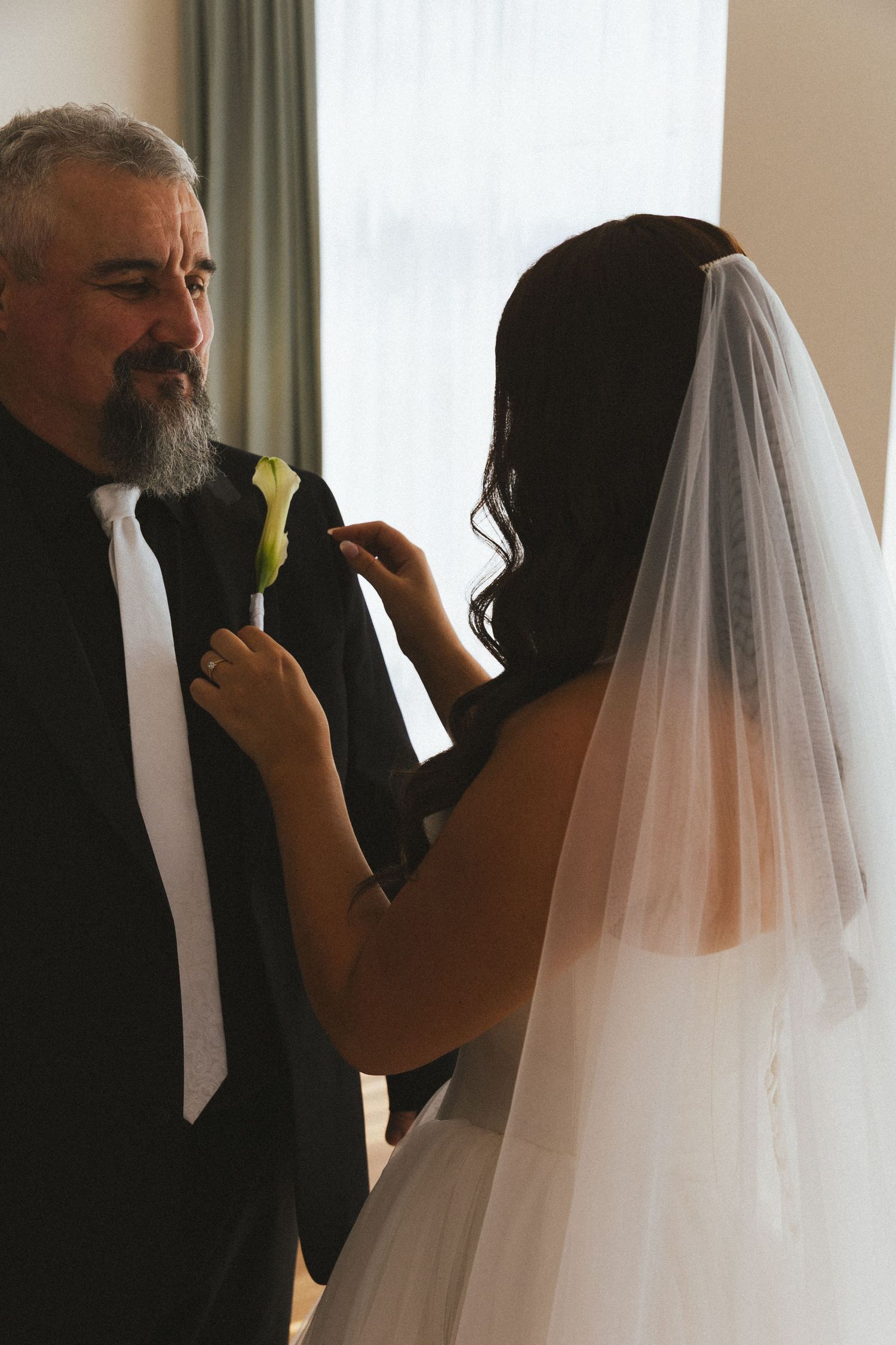 Bride in white veil pinning white calla lily boutonniere on father of the bride in dark suit.