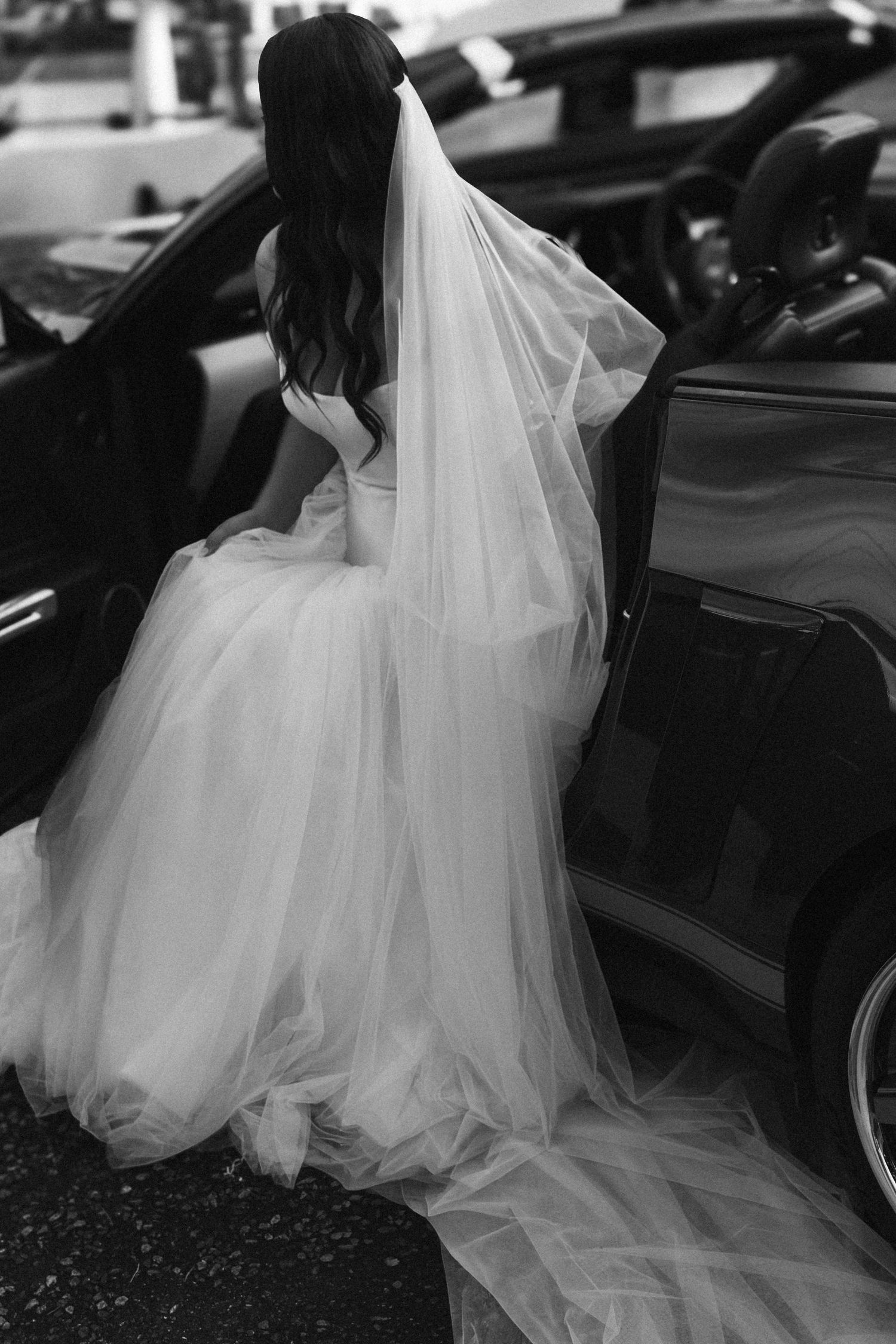 Black and white photo of a bride in a flowing tulle gown and veil stepping into a dark car.