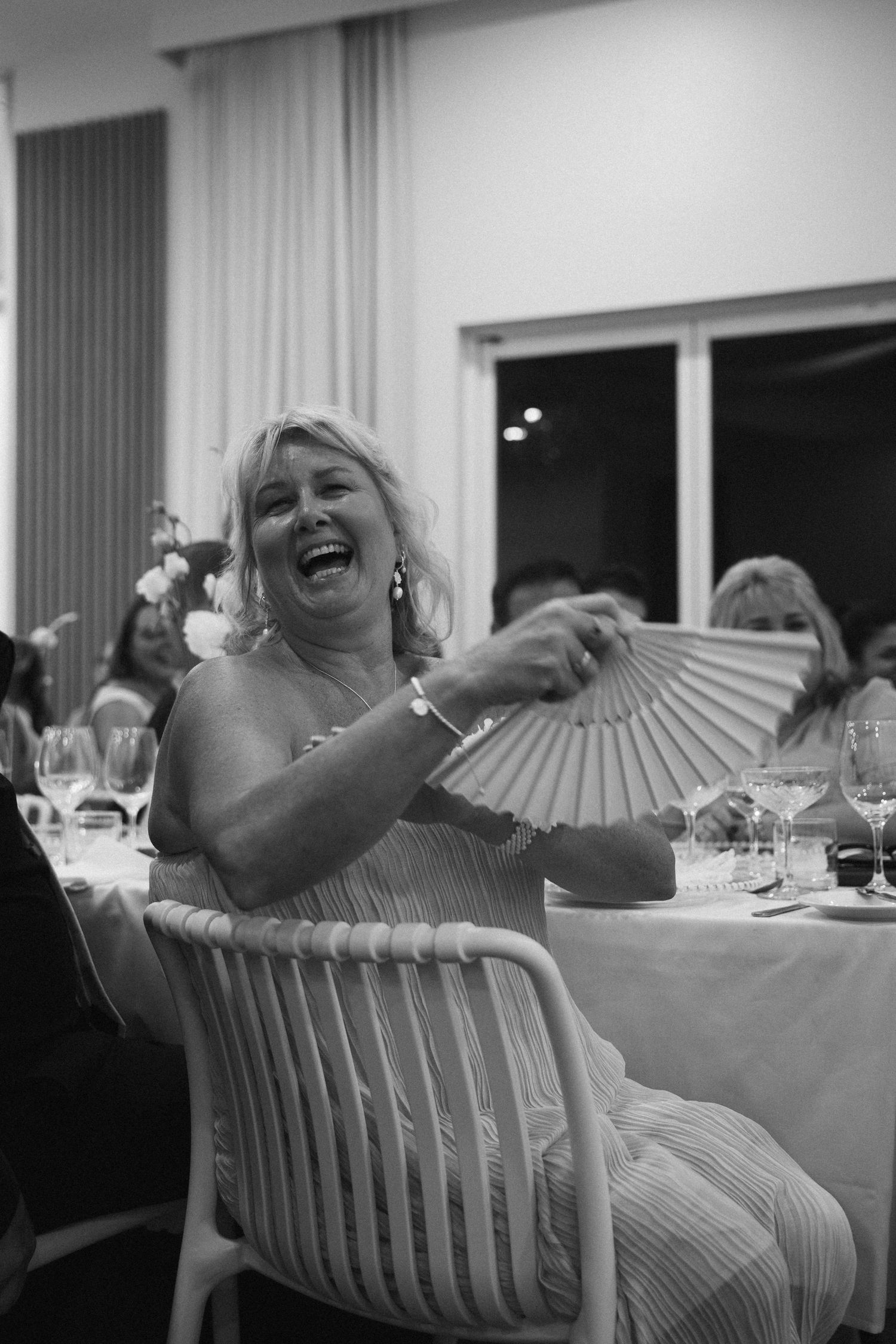 Black and white photo of joyful mother of the bride laughing and holding fan at wedding reception.
