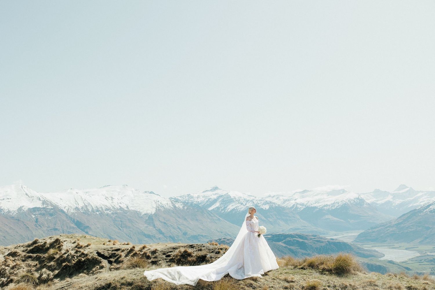 Solitary bride in flowing white gown with long train poses on rocky hilltop with snowy mountain range in Queenstown New Zealand.