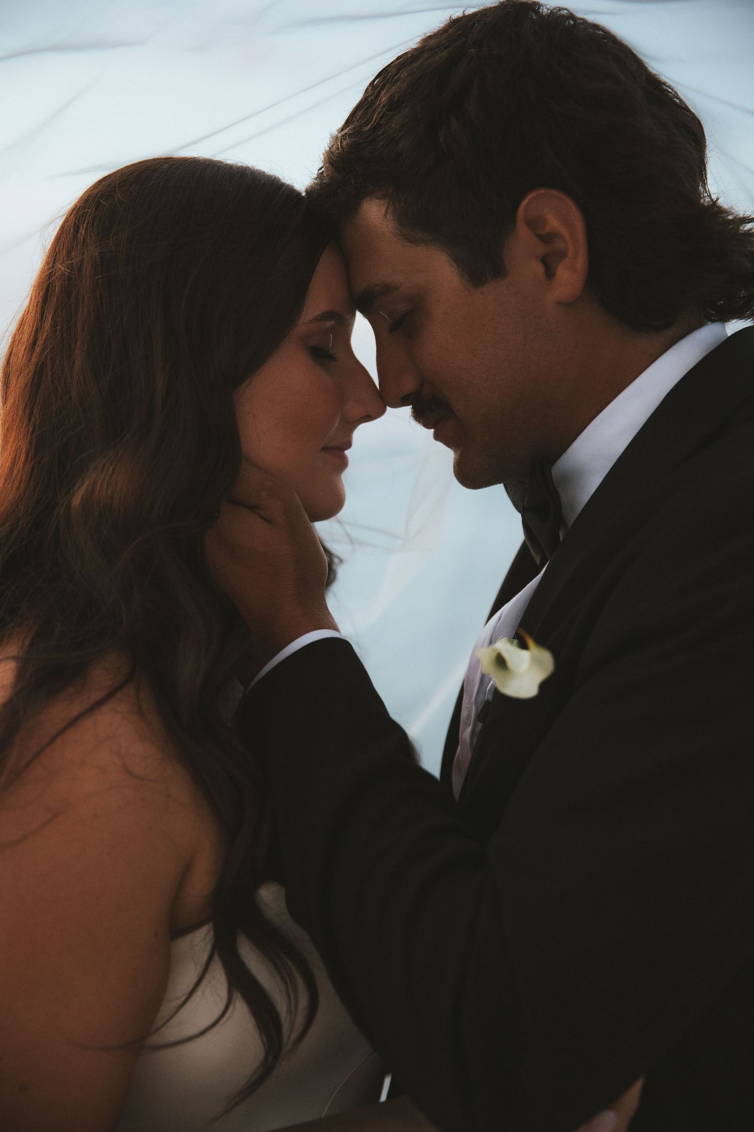 Intimate close-up of bride and groom touching foreheads tenderly under soft glowing light on wedding day.
