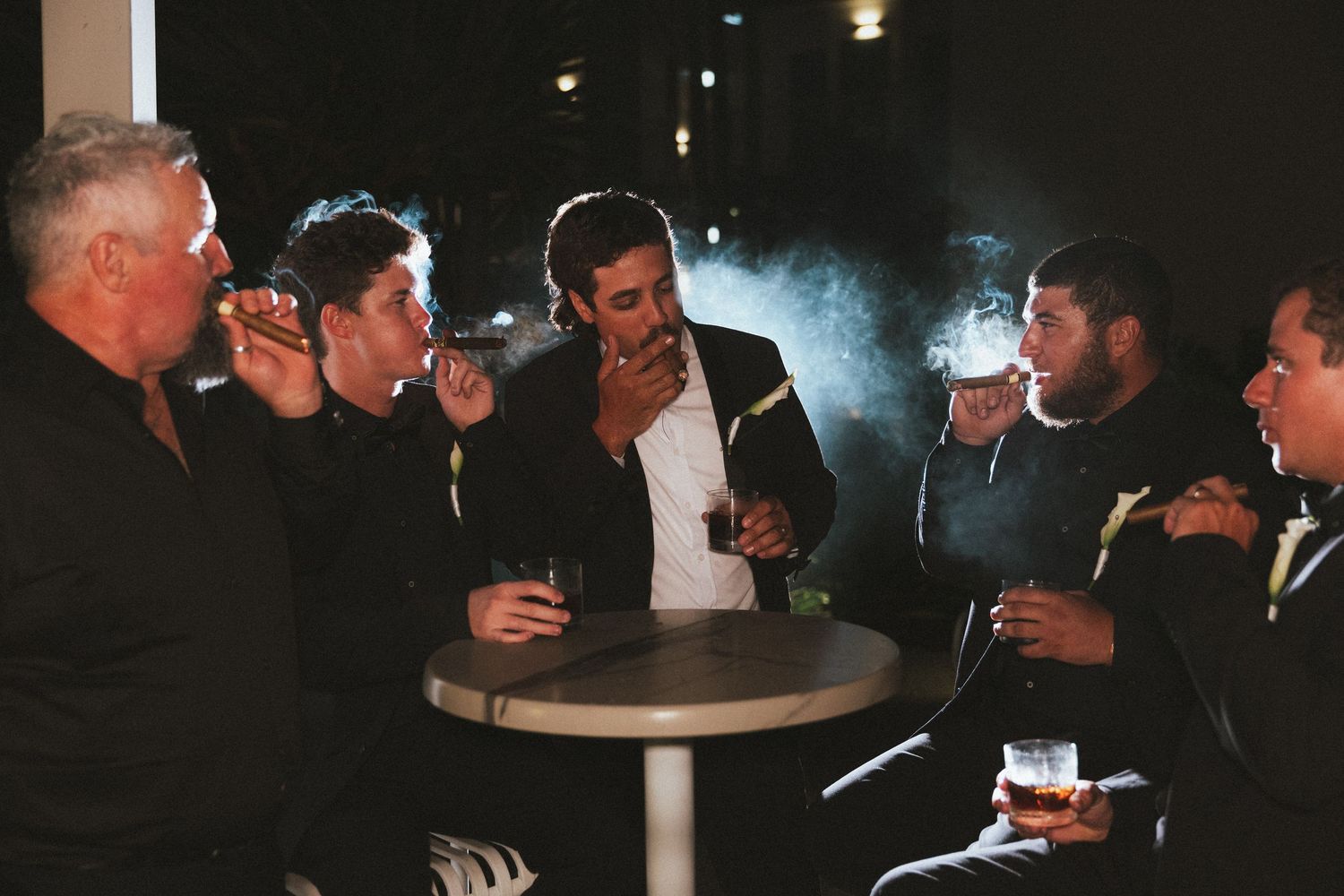 Groomsmen in black suits smoking cigars and drinking whiskey at outdoor evening reception.