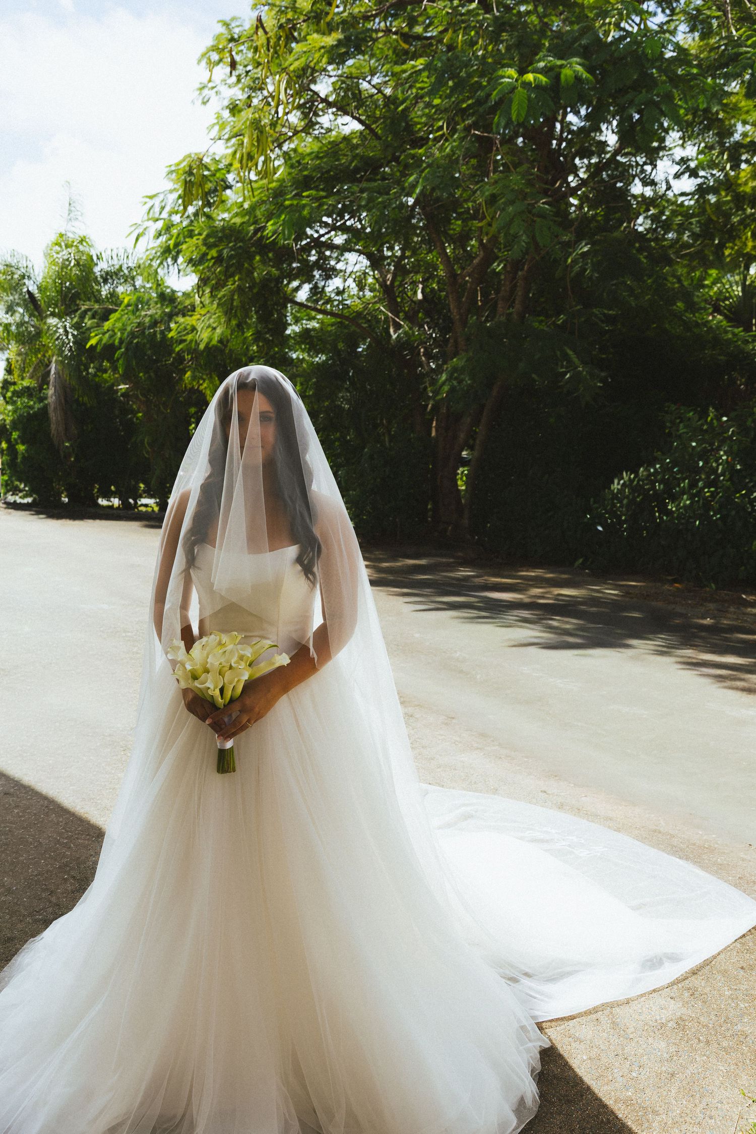 Bride in elegant white Aston Bridal gown with long veil holding bouquet outdoors among green trees.