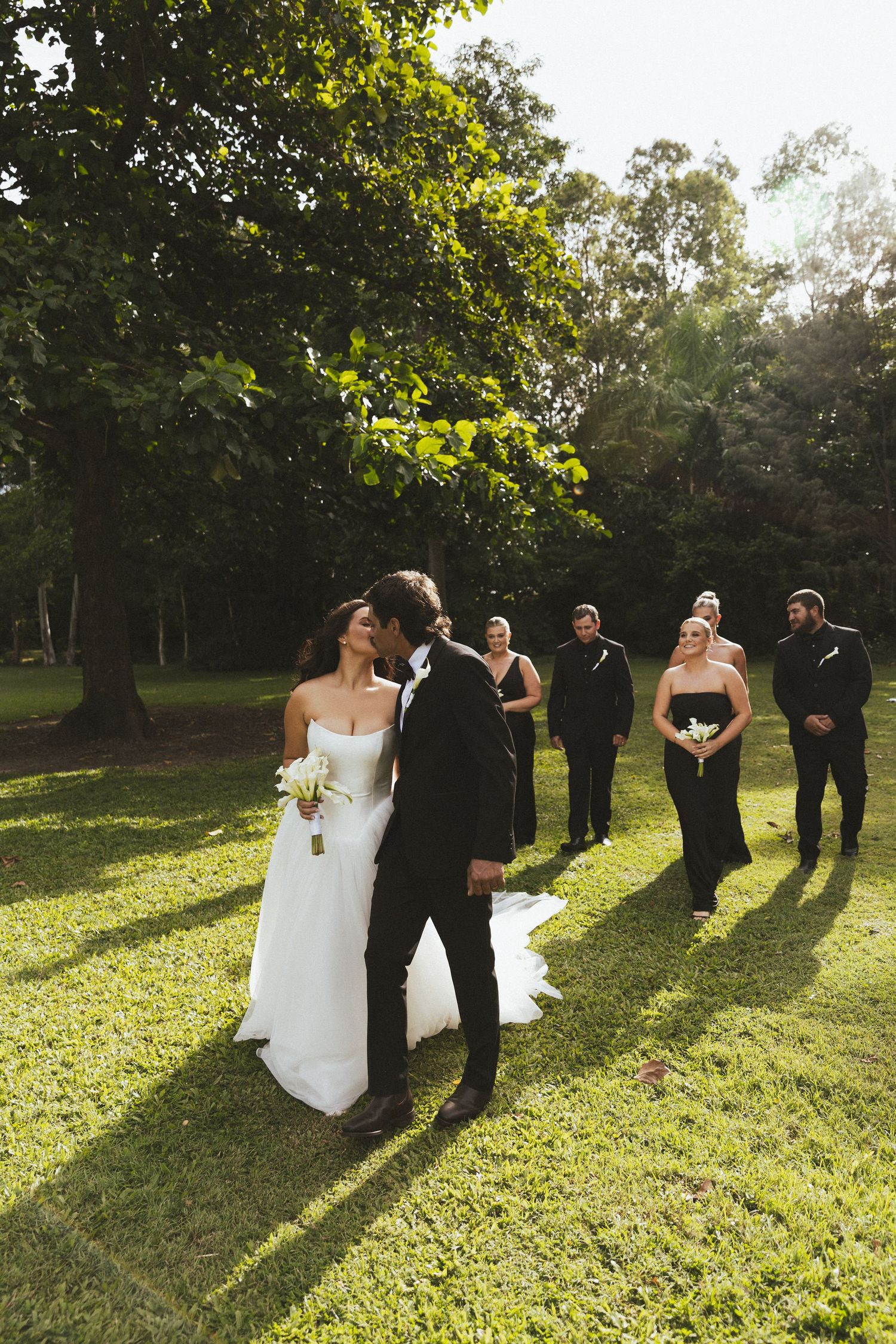 Bride and groom share a romantic moment on green lawn with wedding party in black attire standing behind them.