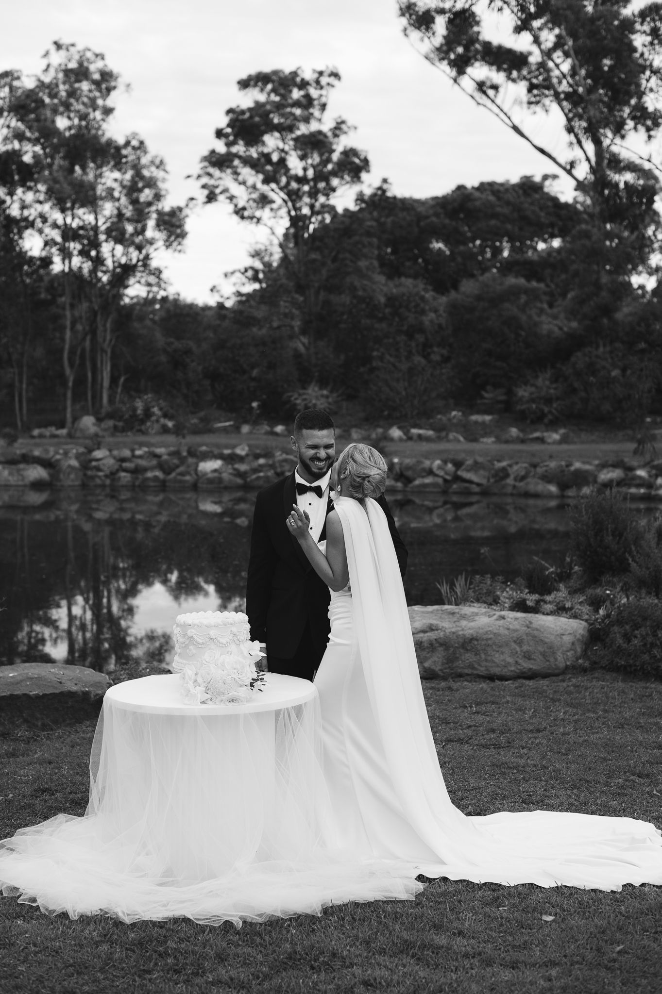 Black and white photo of bride and groom cutting wedding cake by a serene pond surrounded by lush trees.