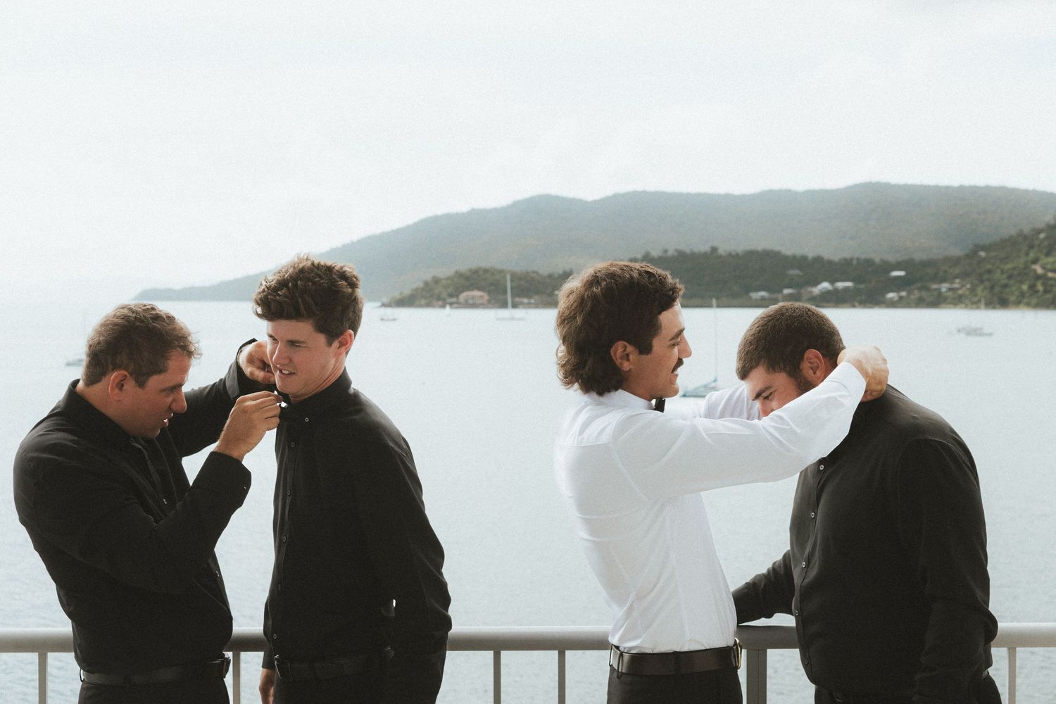 Four groomsmen in black helping each other get dressed on balcony with scenic water view in Whitsundays, Queensland.