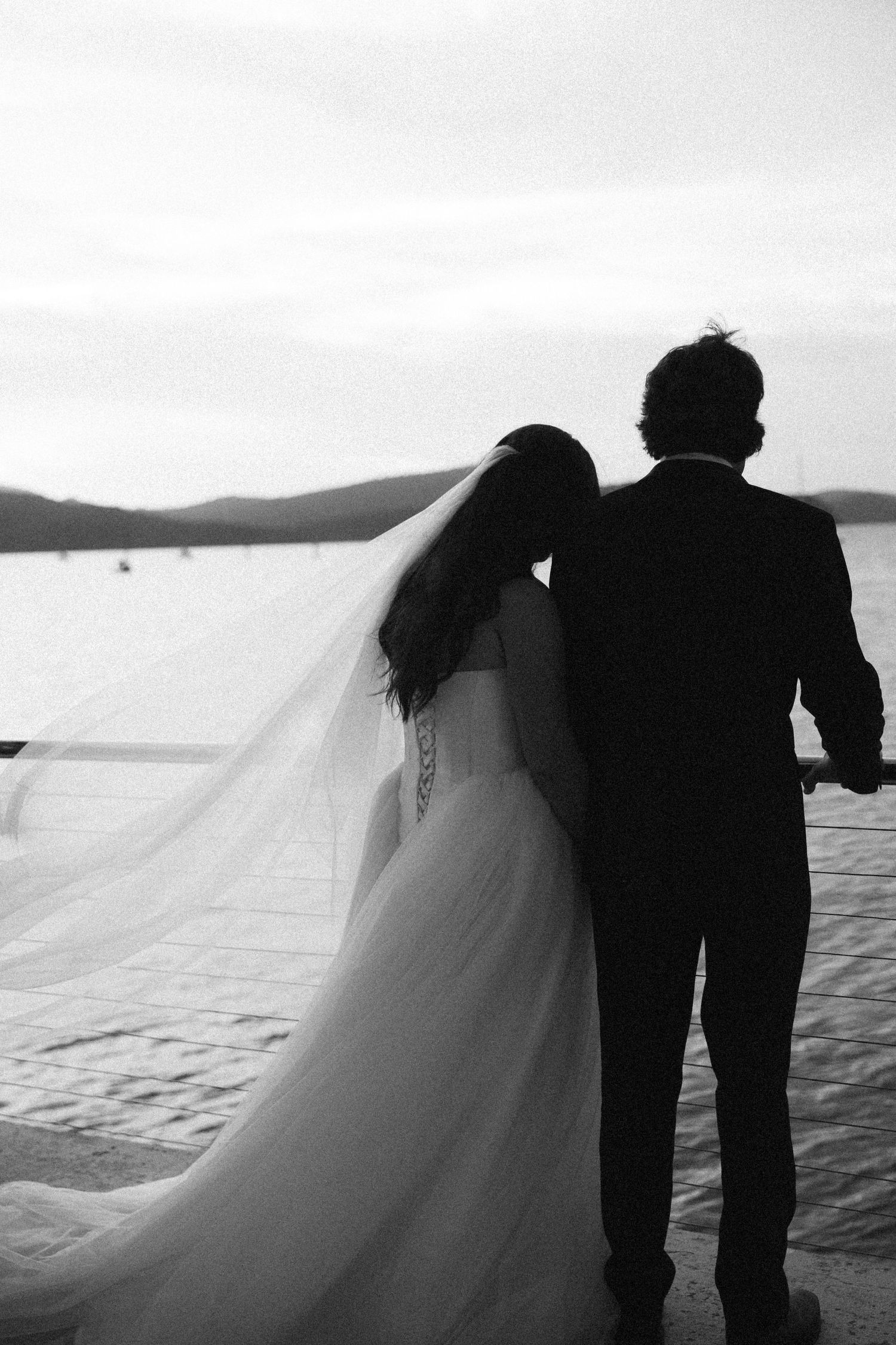 Black and white silhouette of bride and groom walking hand in hand by waterfront at dusk.