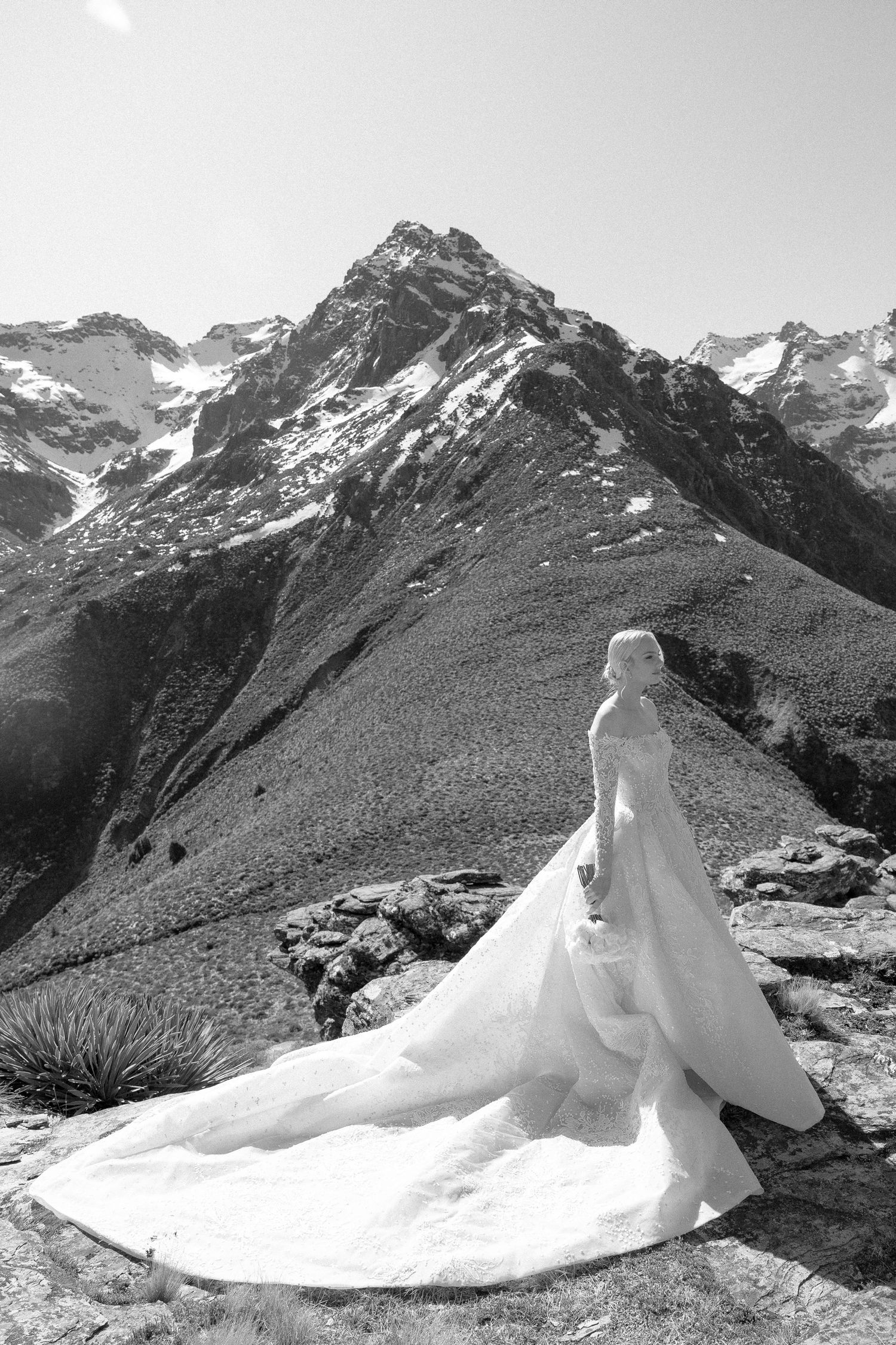 Black and white portrait of bride in ball gown with dramatic train against rugged alpine mountain peak.