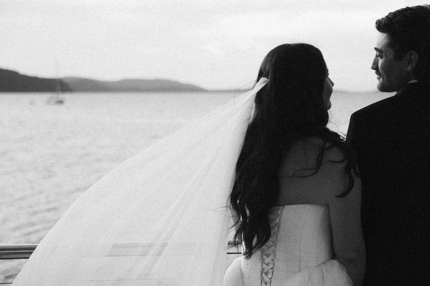 Black and white photo of bride and groom embracing while gazing at ocean view on wedding day.