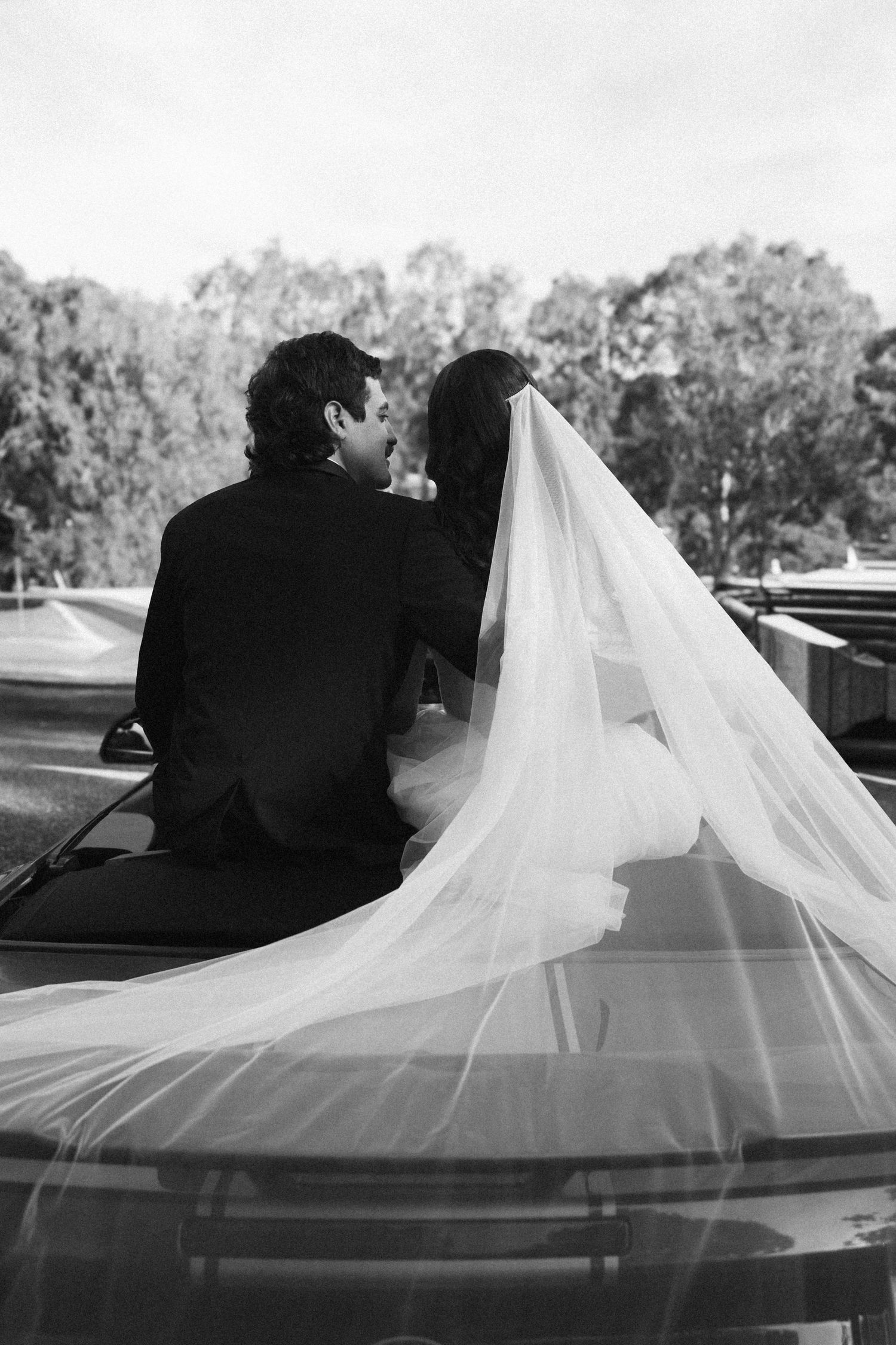 Black and white photo of bride and groom sitting on vintage car hood, brides veil flowing beautifully.