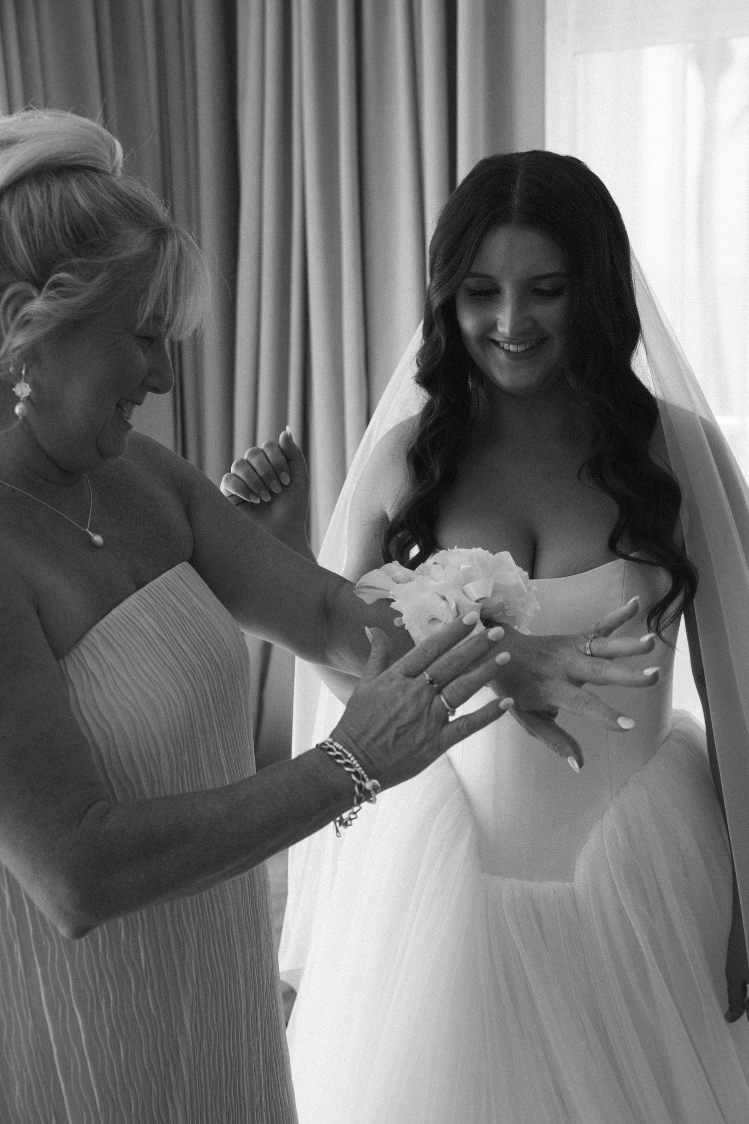 Black and white photo of mother helping radiant dark-haired bride with bouquet while getting ready near a window.