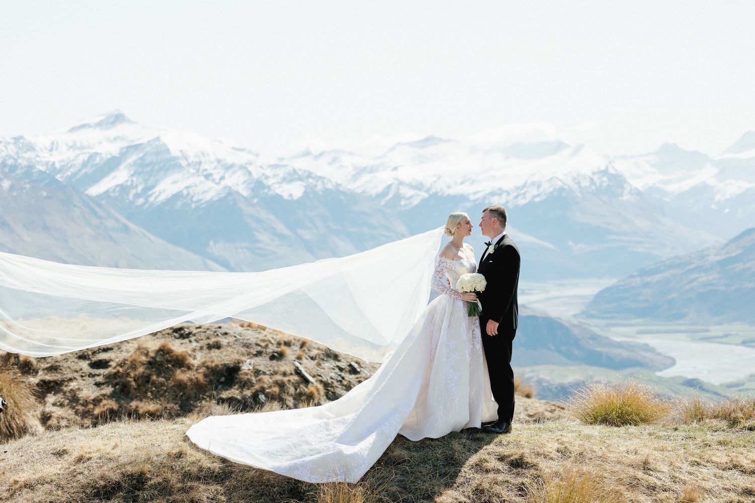 Bride and groom embrace on mountain summit with stunning snow-capped peaks and valley views behind them in Queenstown, New Zealand.