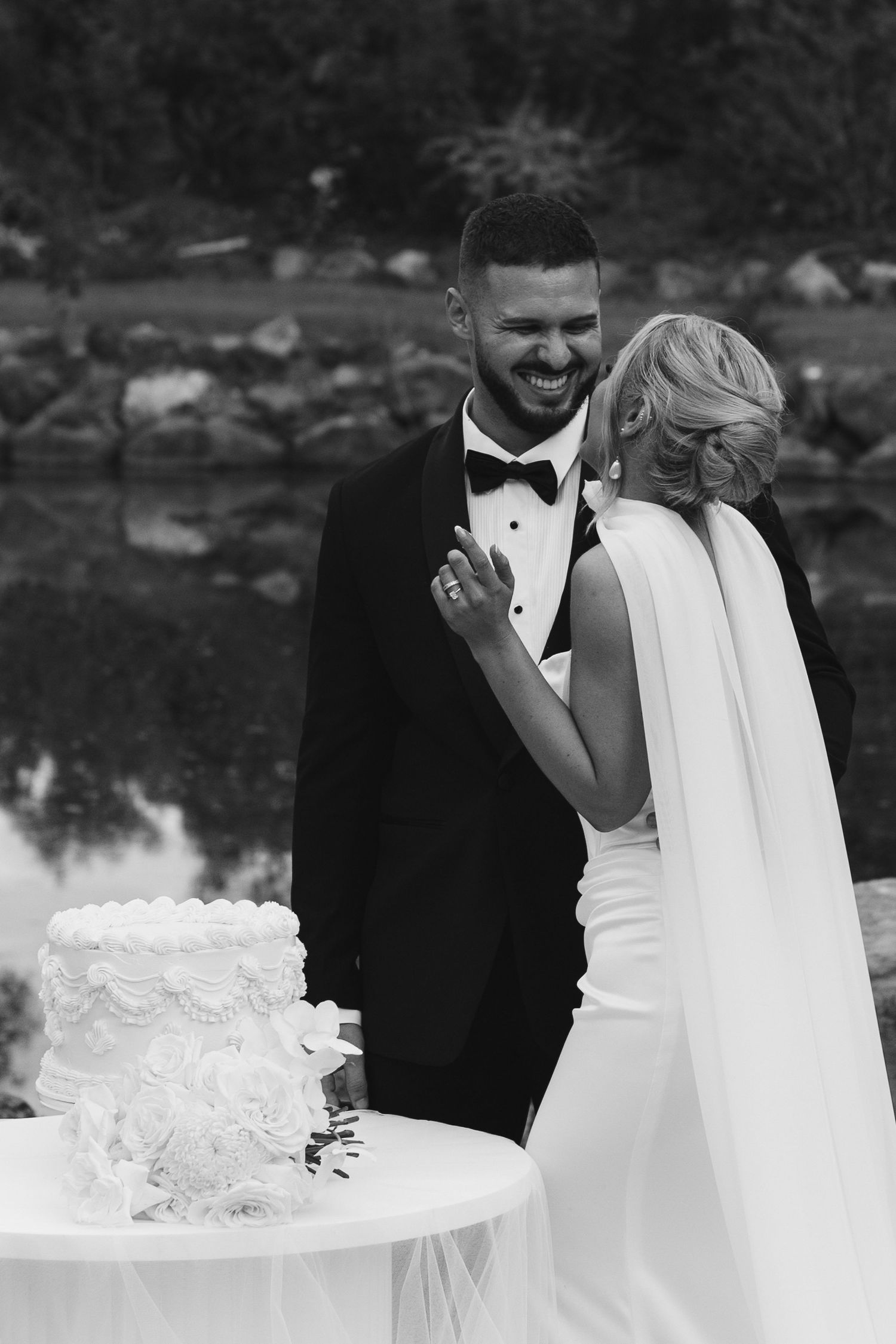 Black and white photo of a laughing groom in a tuxedo and bride in a white gown cutting their wedding cake outdoors.