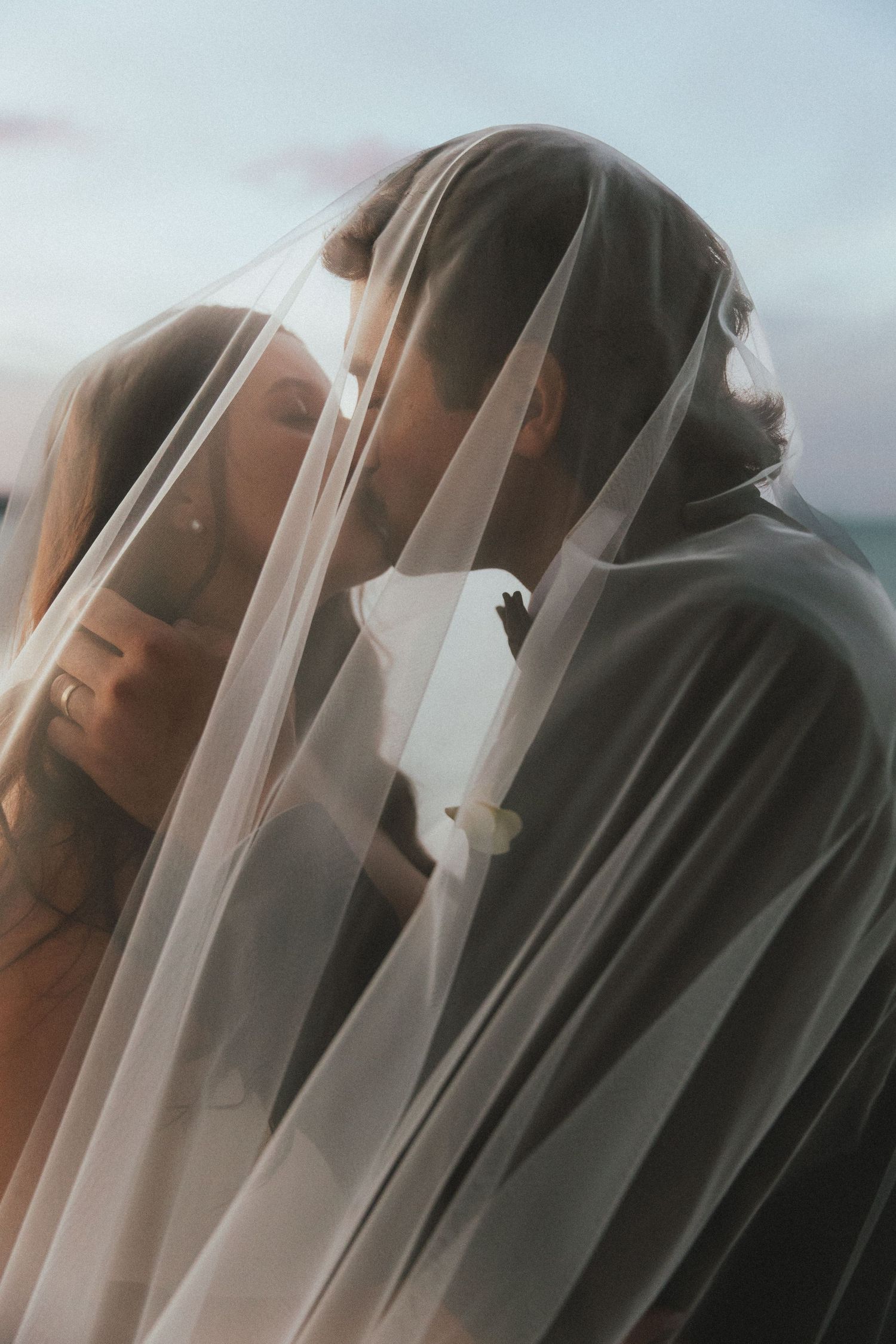 Bride and groom sharing a romantic kiss under a sheer wedding veil at dusk.