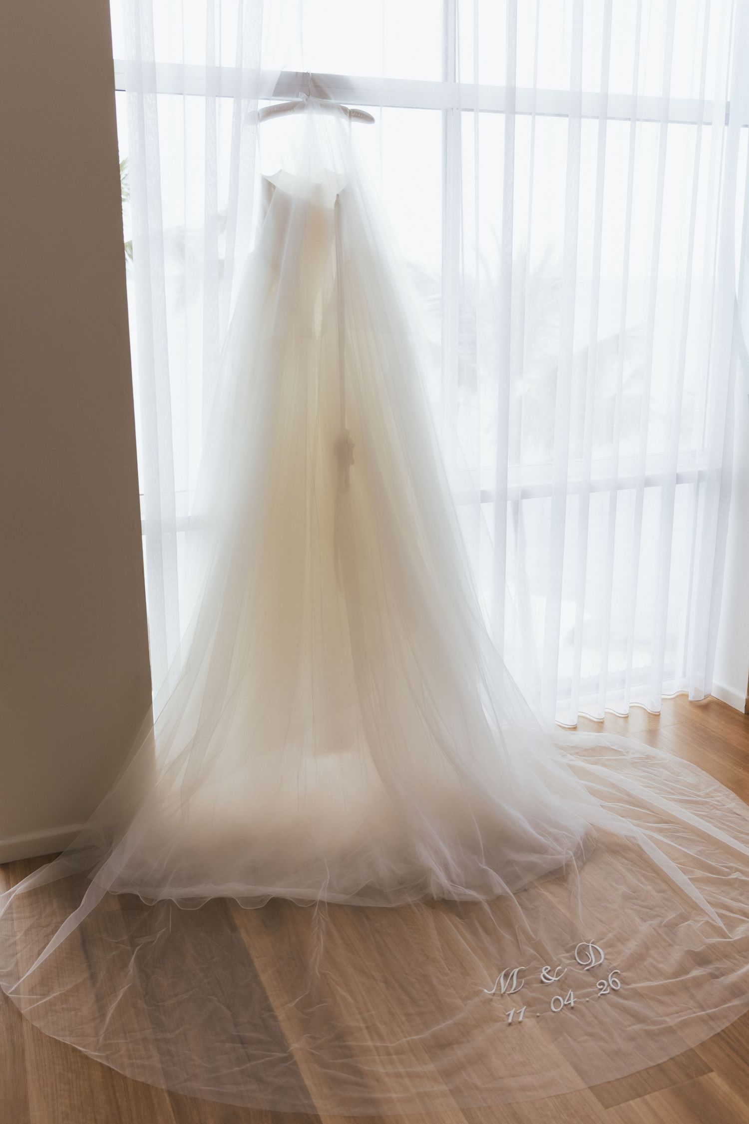 Elegant white tulle Aston Bridal wedding gown with monogrammed veil displayed hanging by a bright window.