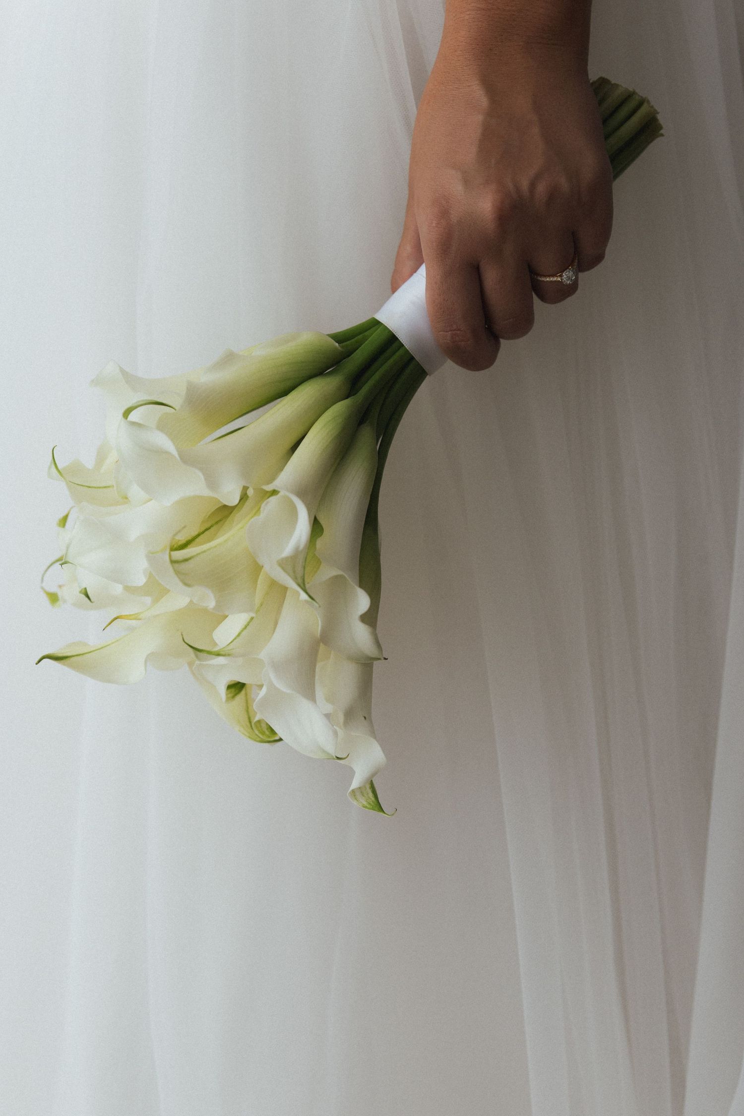 Bride holding elegant white calla lily wedding bouquet wrapped with white ribbon, wearing diamond engagement ring.