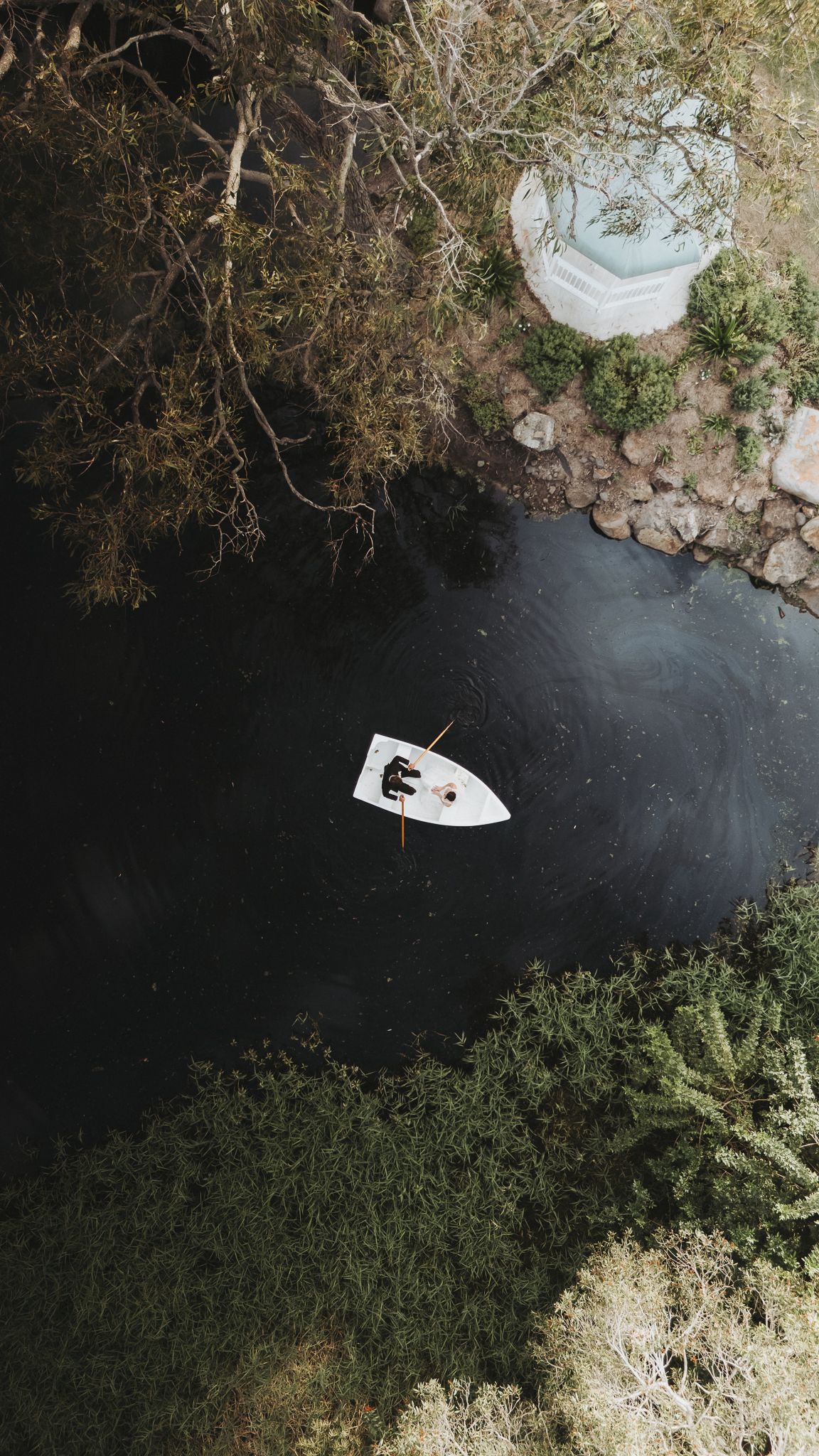 Aerial drone view of a white boat with a person rowing on a dark calm river surrounded by lush green trees.