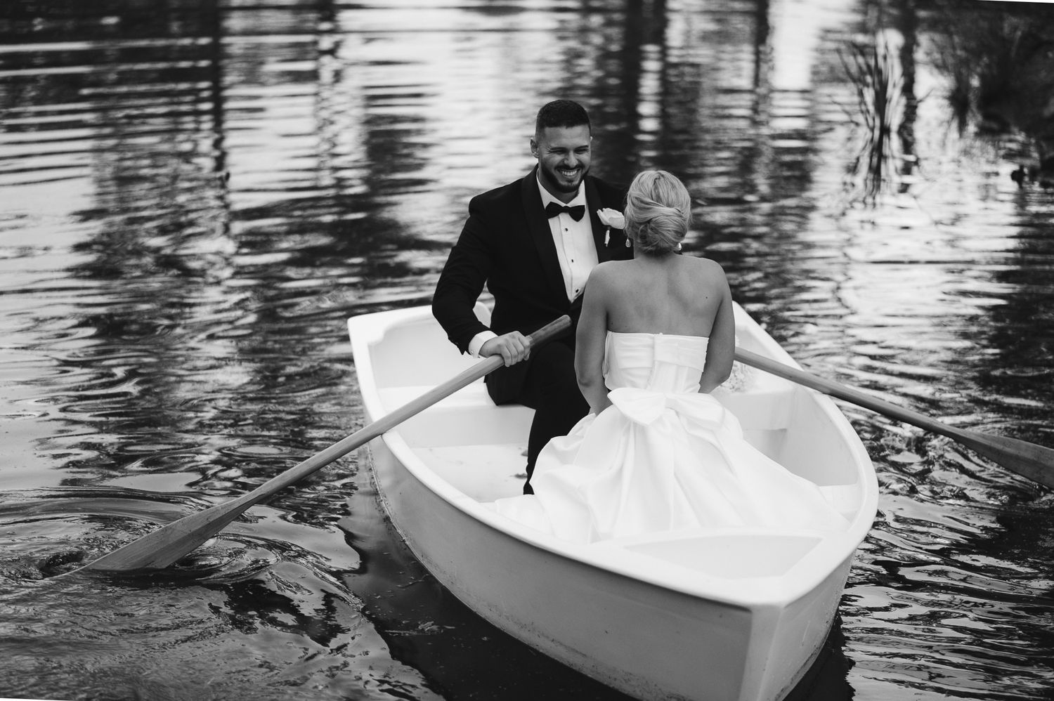 Groom rows bride in a white boat on a lake in an elegant black and white wedding portrait.