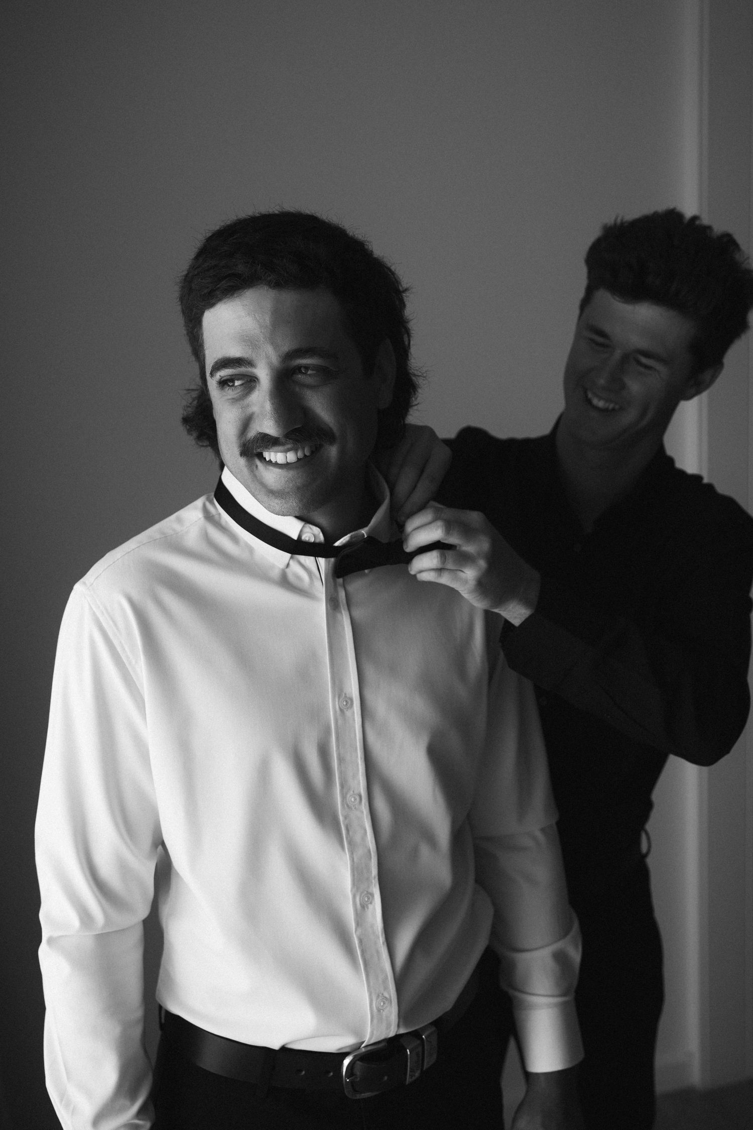 Black and white photo of smiling groom getting bow tie adjusted by groomsman before wedding.