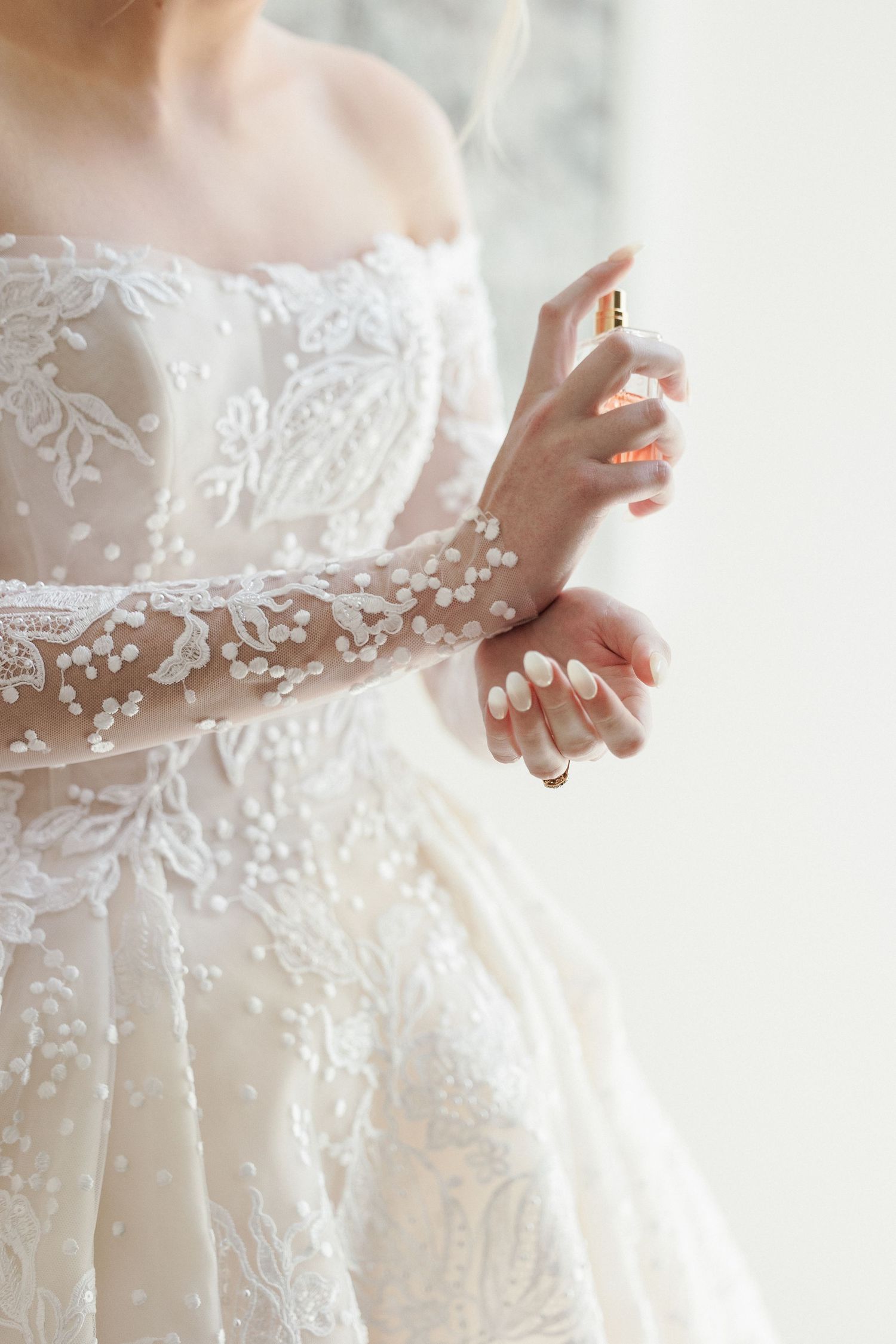 Close-up of bride in intricate lace off-shoulder Steven Khalil gown applying Chanel perfume to her wrist.