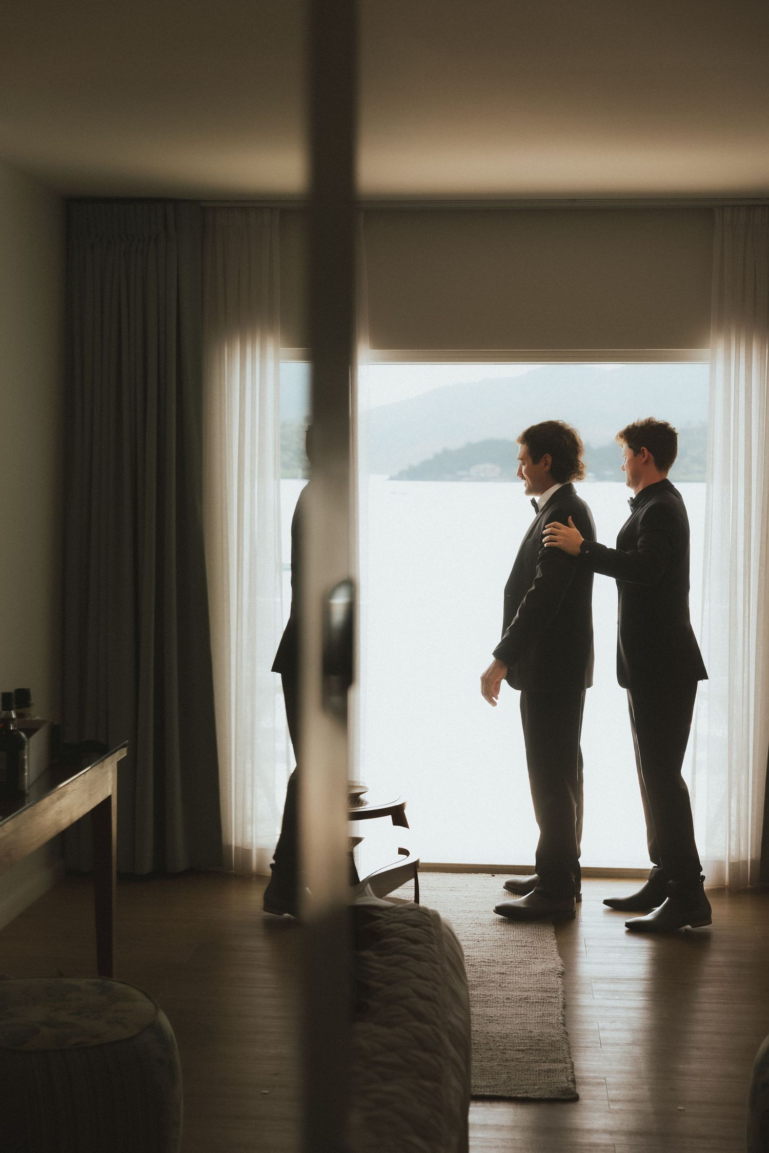 Two men in black suits stand silhouetted by a bright window with a scenic water view while getting ready.