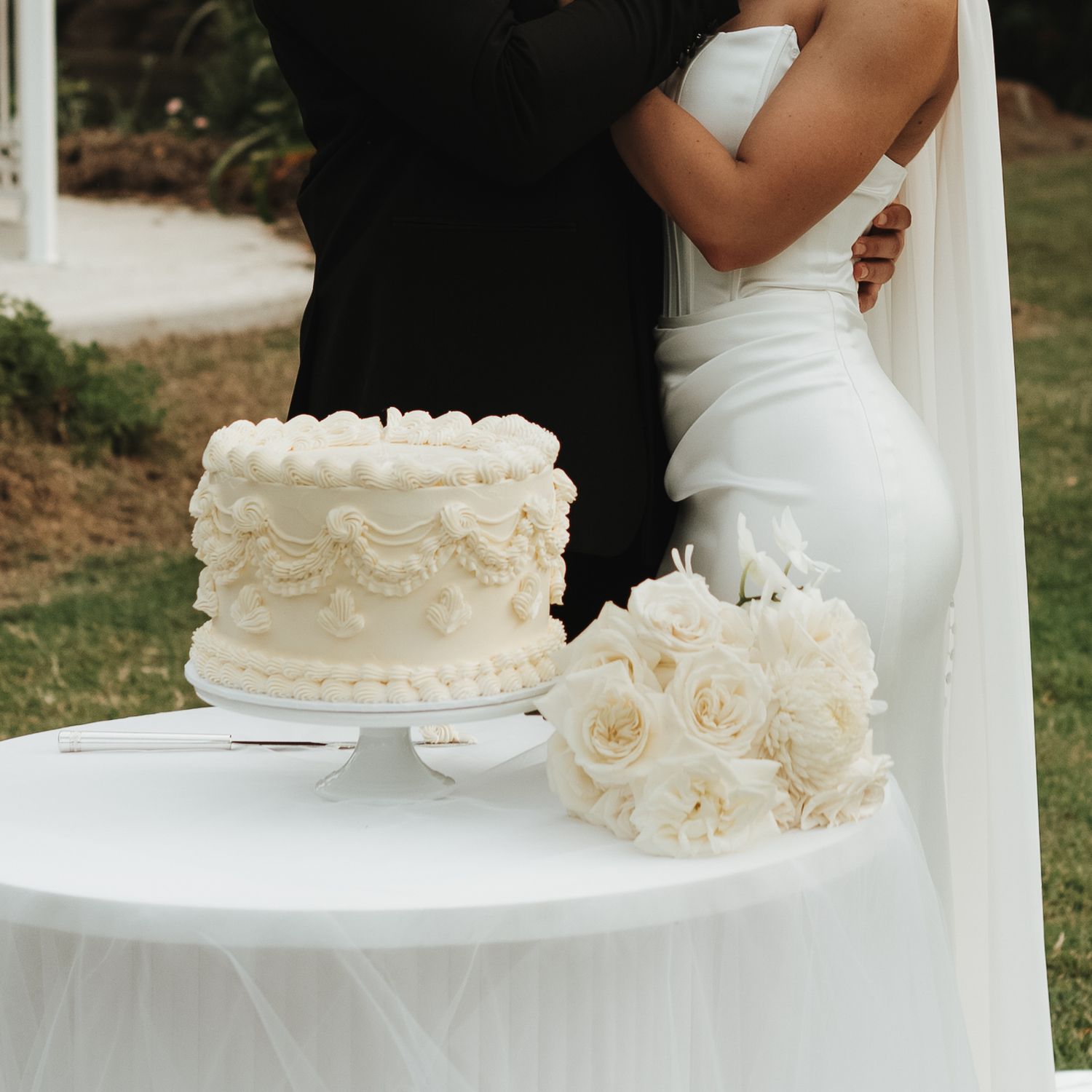 Couple embracing behind a elegant white wedding cake and rose bouquet at an outdoor garden reception.