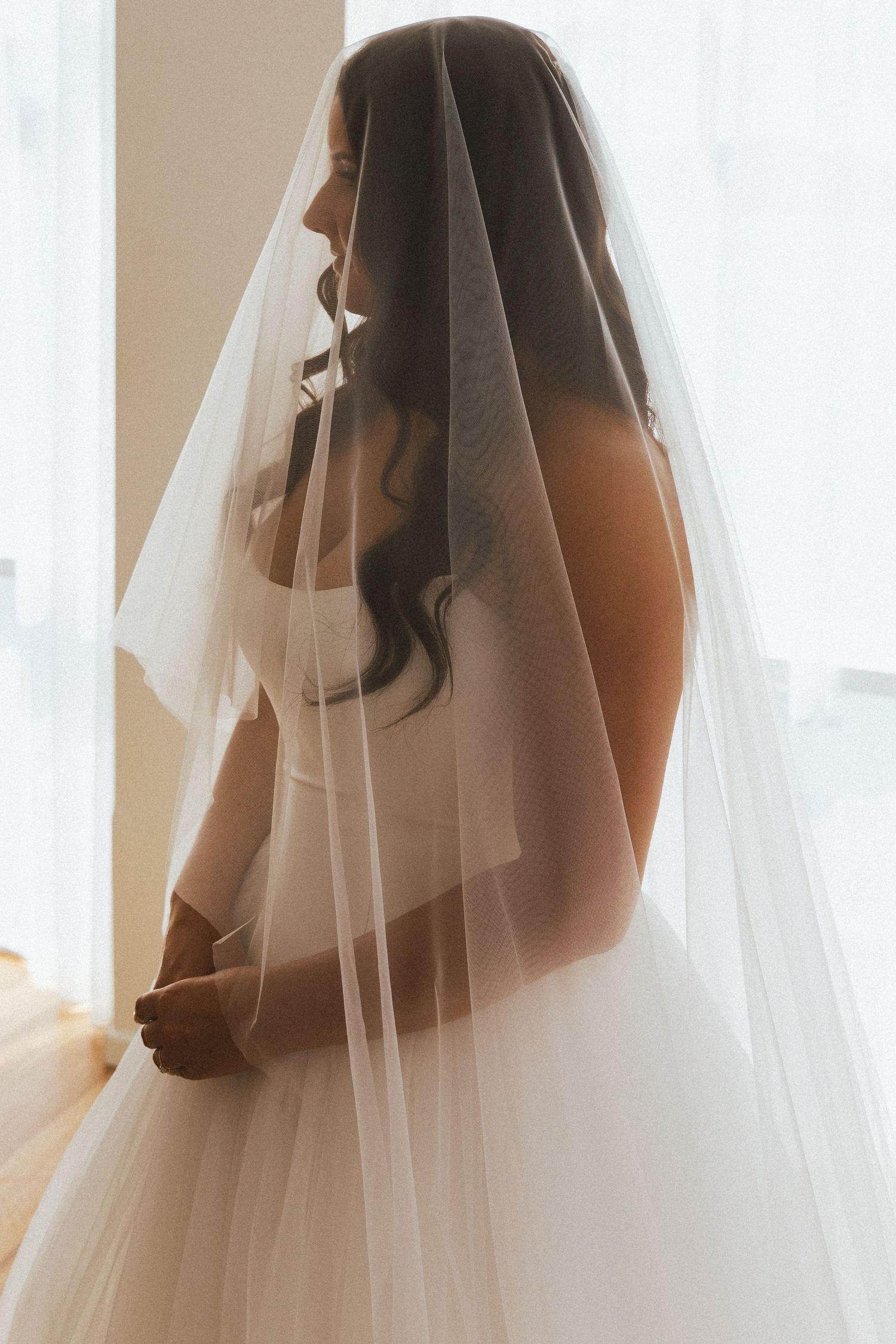 Beautiful bride in white strapless Aston Bridal gown with flowing cathedral veil gazing out bright window.