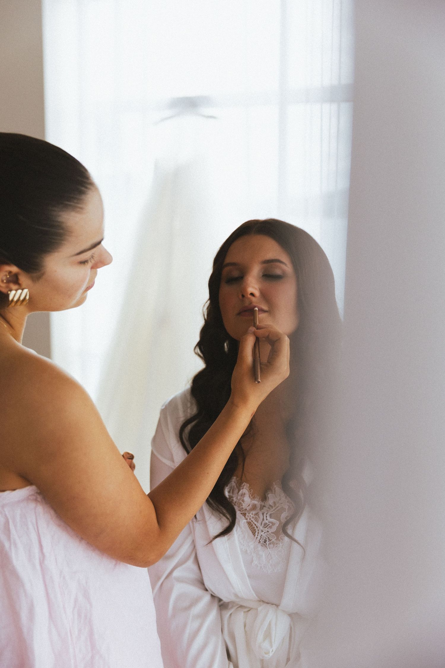 Makeup artist applying lipstick to dark-haired bride in white robe during wedding morning preparations.