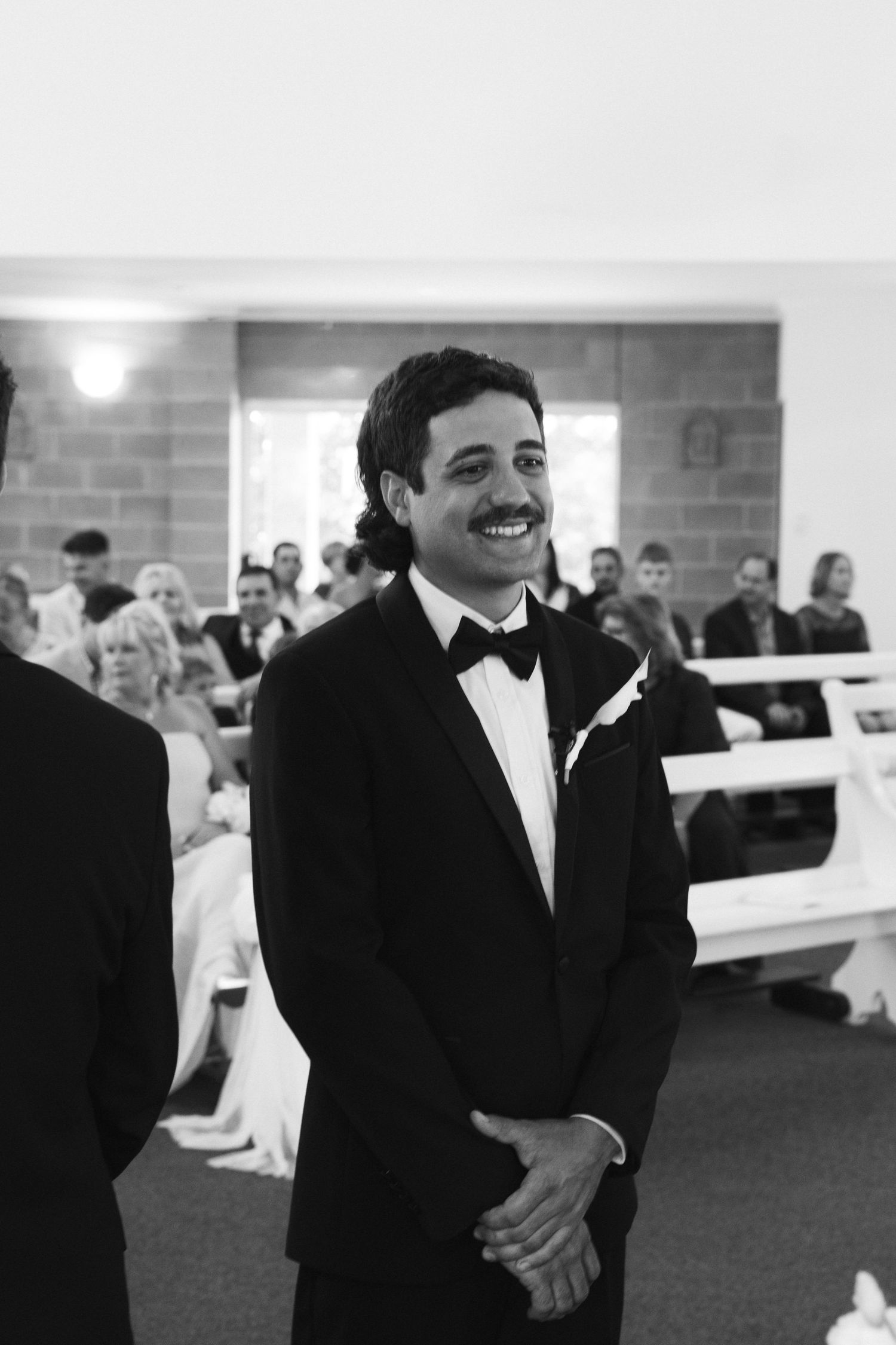 Black and white photo of smiling groom in tuxedo waiting at church altar on wedding day.