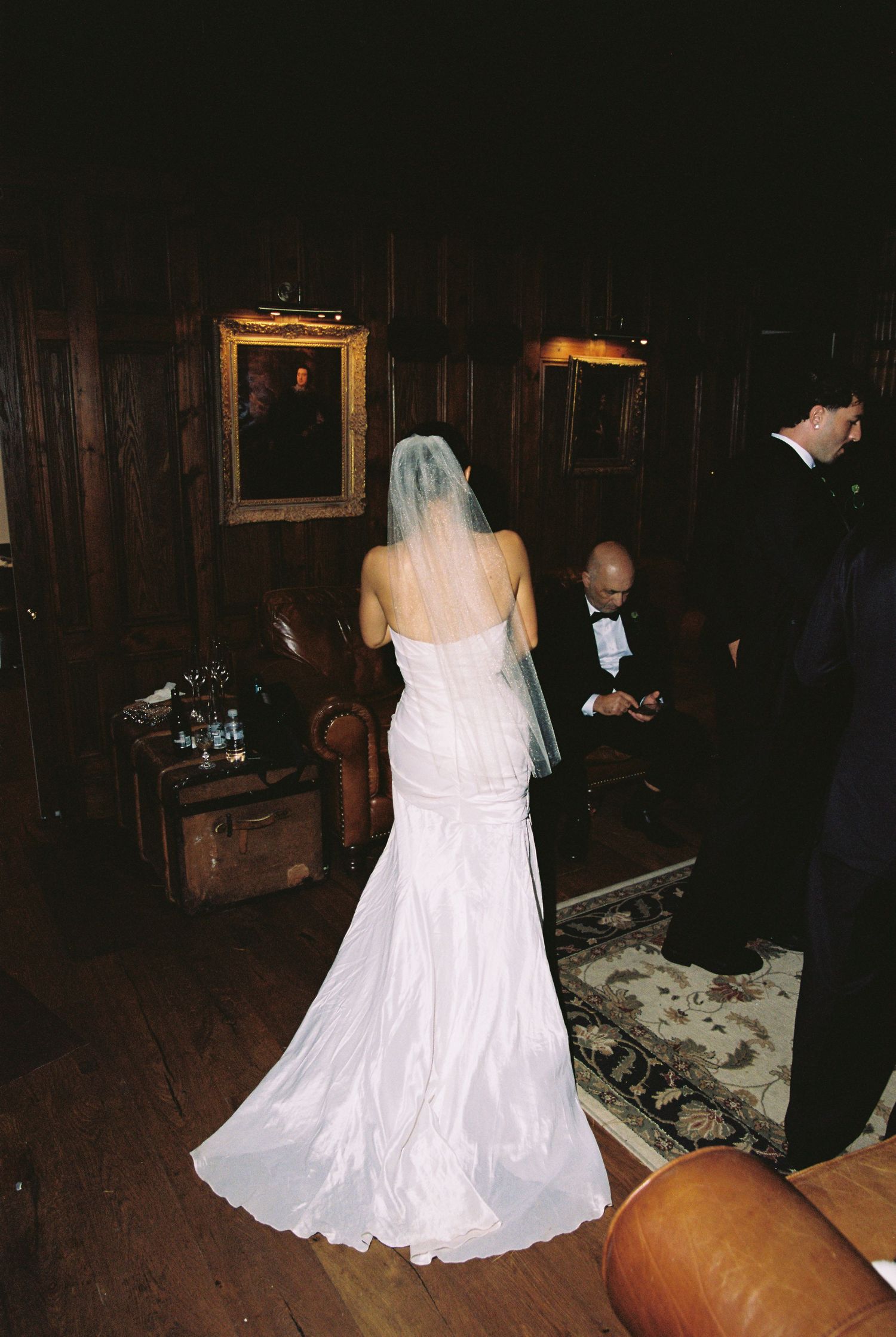 Bride in white gown with veil standing in a dark wood-paneled room with portraits.