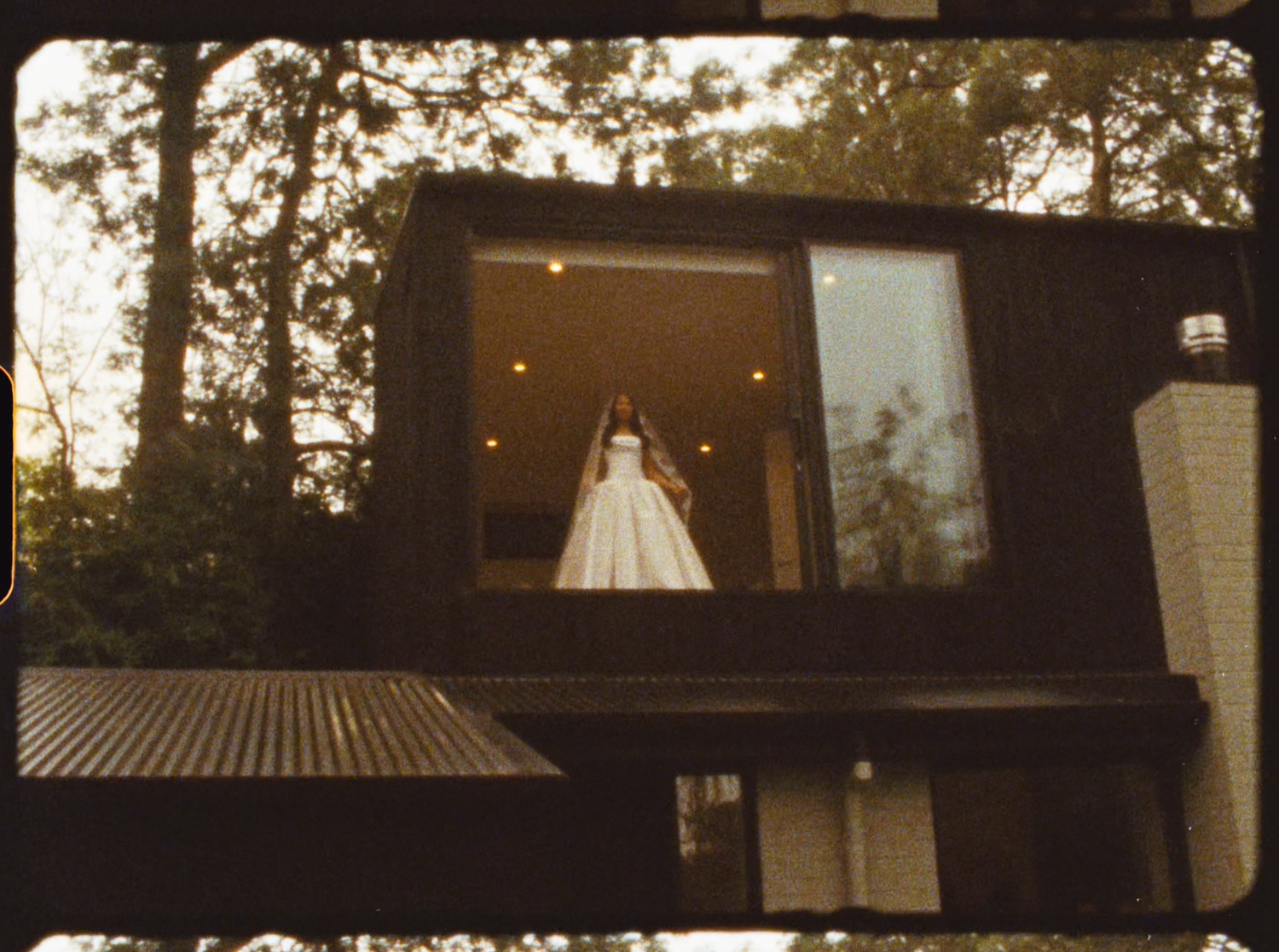 Bride in white ball gown standing in a large window of a modern dark wooden building.