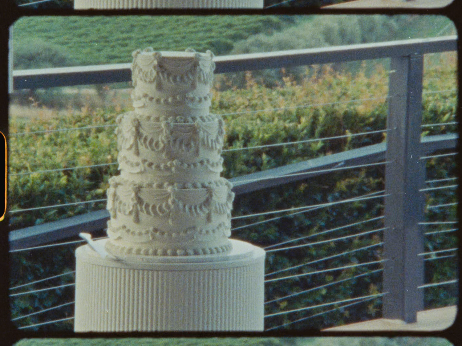 Elegant four-tier white wedding cake with floral details displayed on a stand outdoors.