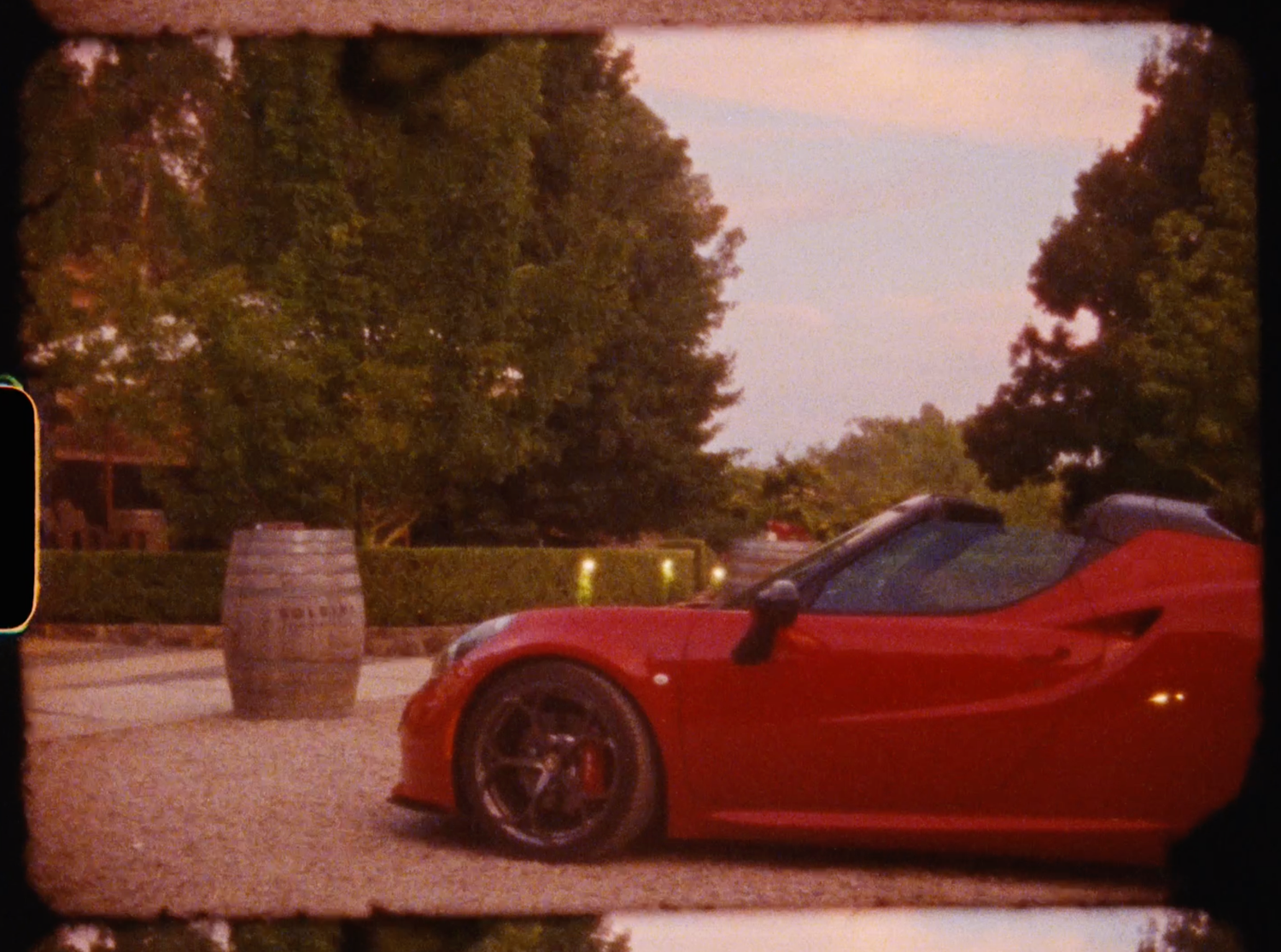 Red Ferrari sports car parked beside a wooden barrel in a scenic outdoor venue at dusk.