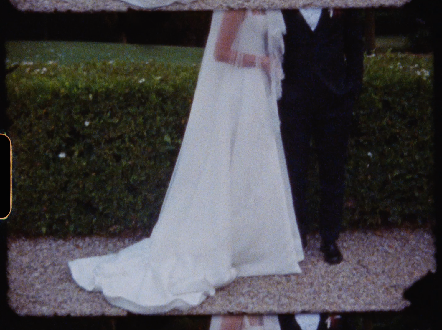 Bride in elegant white wedding gown with long train standing beside groom in dark suit outdoors.