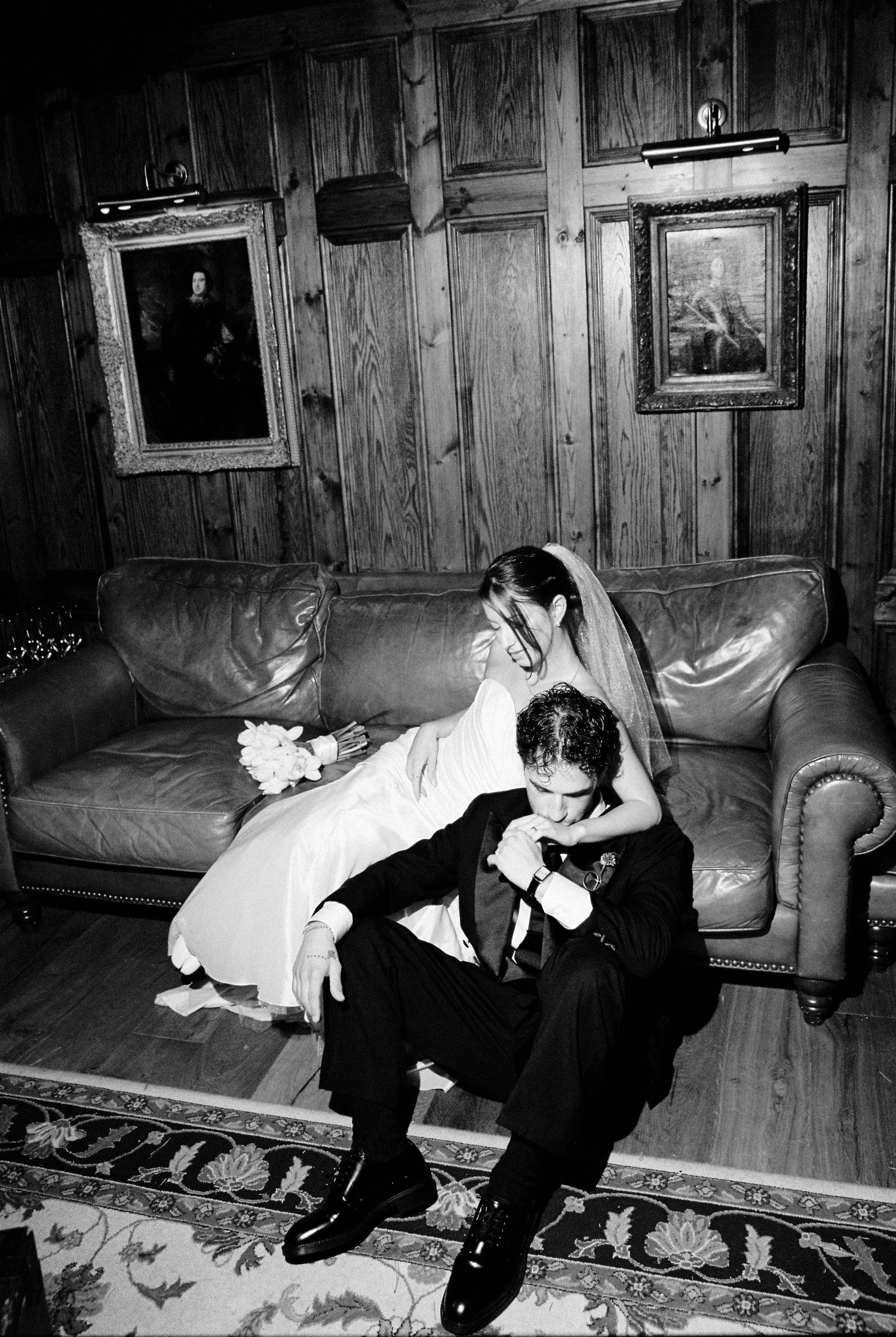 Bride and groom share an intimate moment seated on a leather sofa in a classic wood-paneled room.
