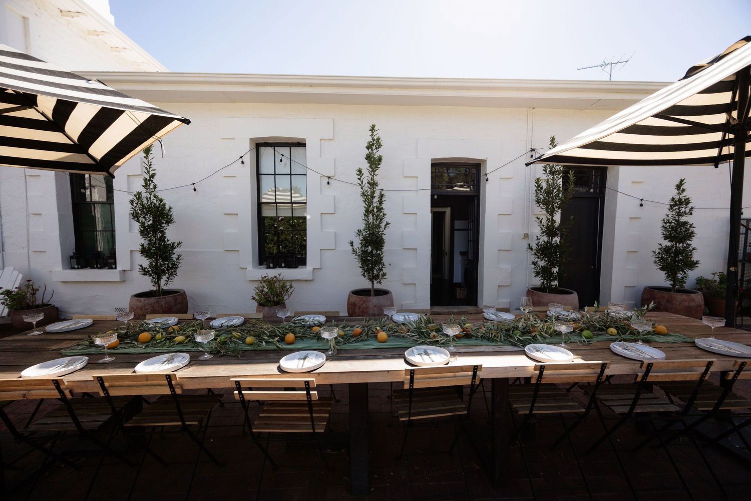 Elegant outdoor wedding reception with long wooden table, striped umbrellas, and lush greenery against white building.