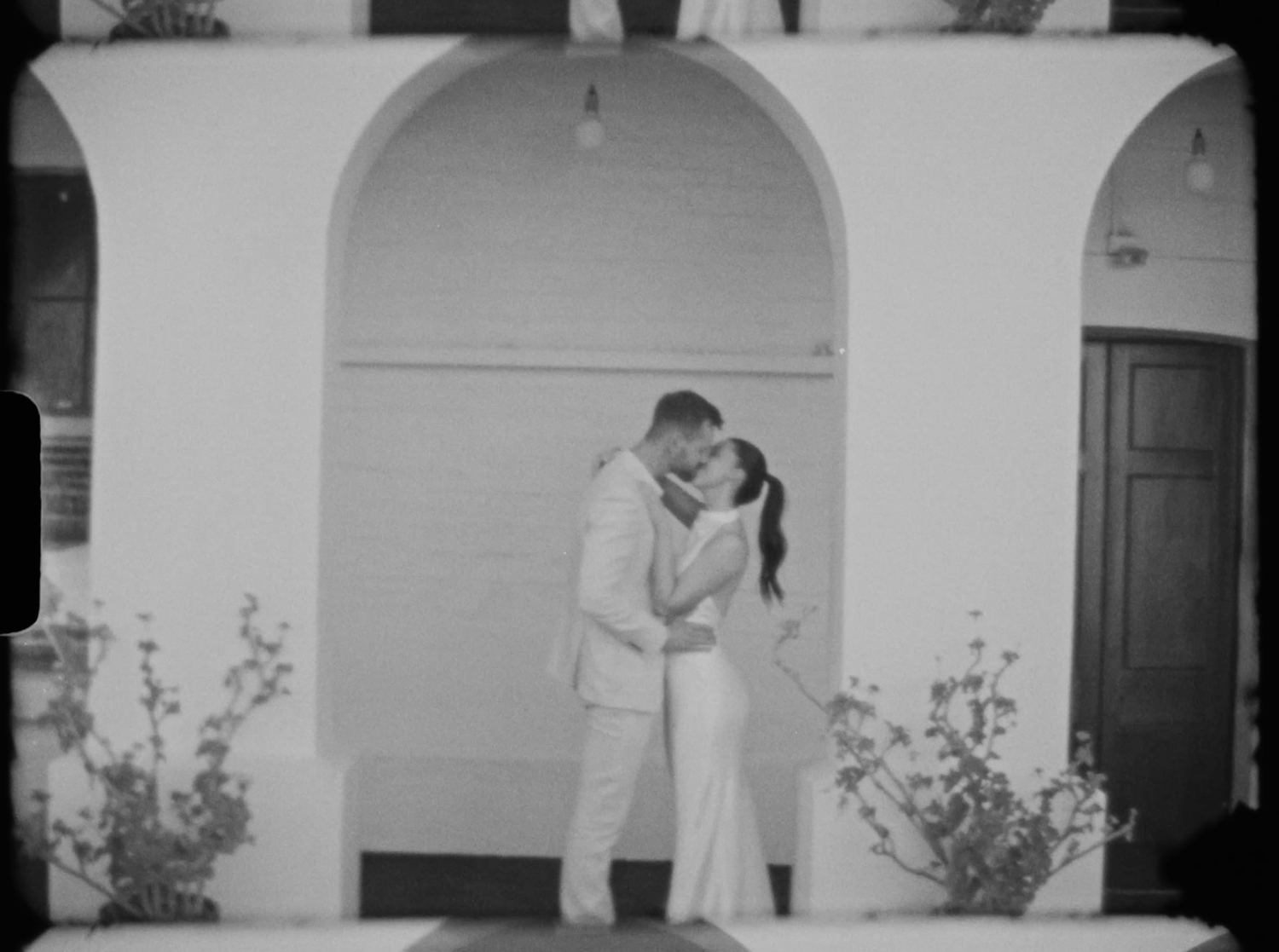 Couple sharing a romantic kiss under a white arched doorway in black and white wedding photo.