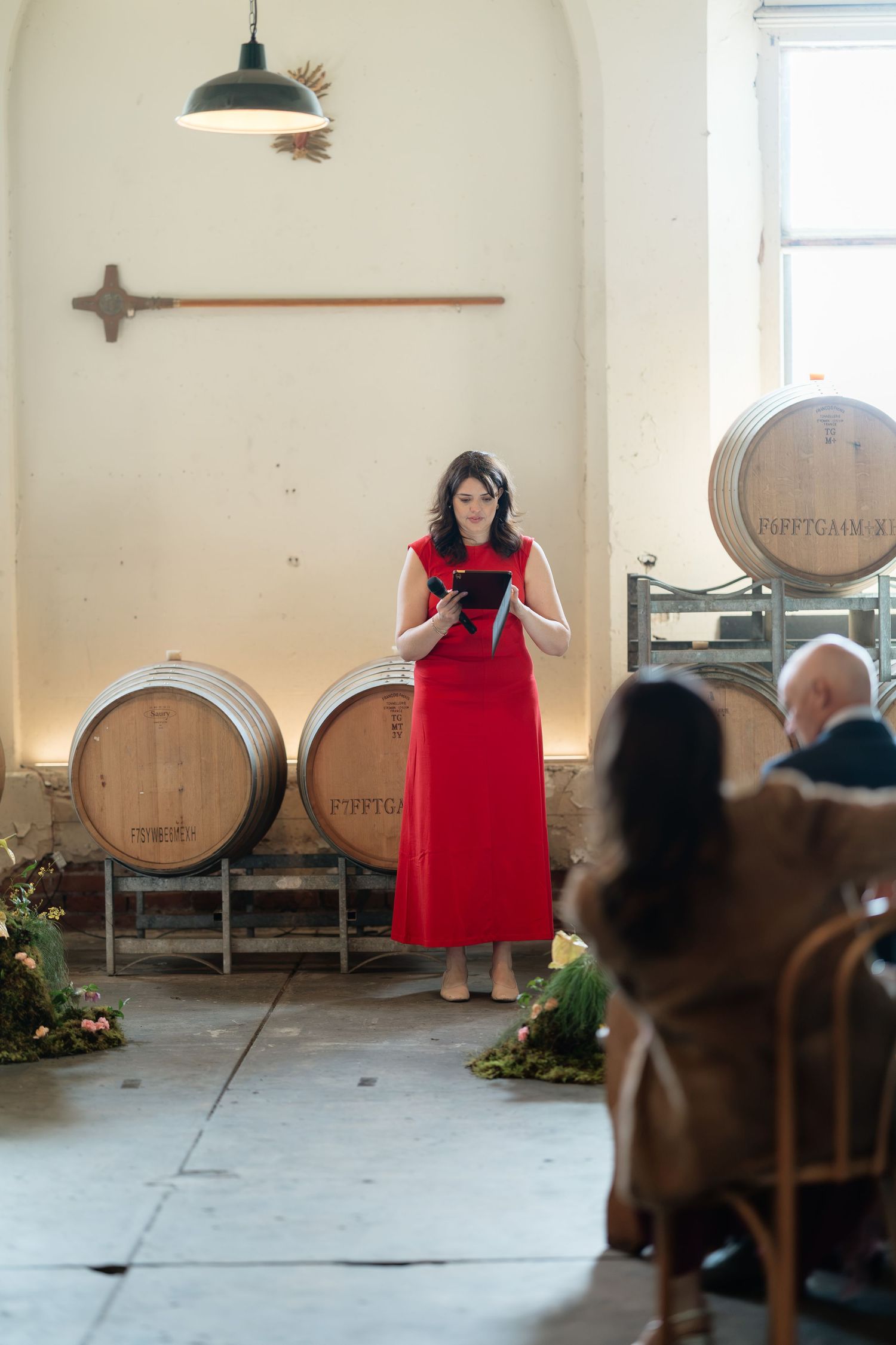 Woman in red dress officiating a wedding ceremony in a rustic winery with oak barrels.
