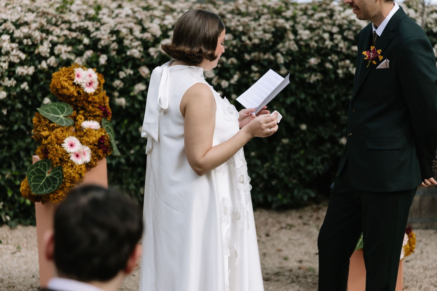 Bride reads vows to groom during outdoor wedding ceremony with floral arch and lush white blooms in background.